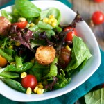 A fresh Farmer's Market Salad with Mustard Vinaigrette in a white bowl, featuring mixed greens, cherry tomatoes, corn, and roasted potatoes, placed on a green cloth napkin with extra tomatoes and spinach leaves scattered around.