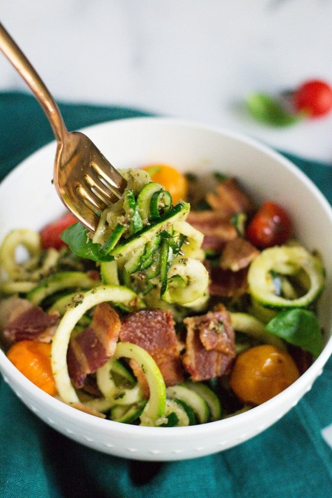 A bowl of zucchini noodles with bacon and cherry tomatoes with a fork full of zoodles.