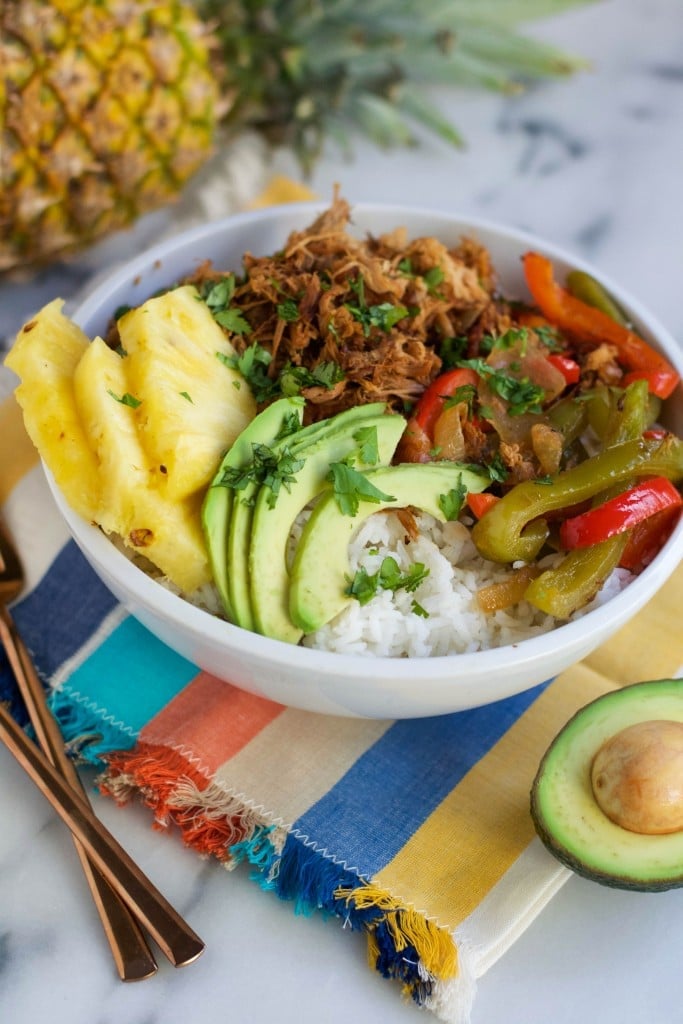 A bowl of slow cooker pork, pineapple, avocado, rice, and roasted peppers with a pineapple and avocado in the background.