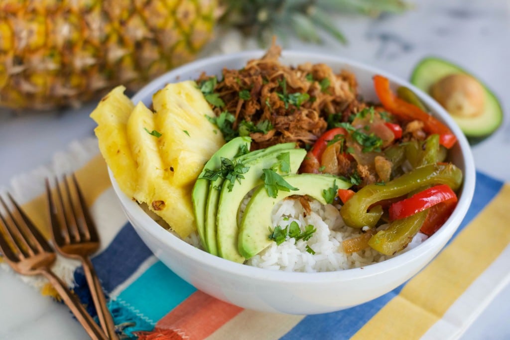 A bowl of slow cooker shredded pork, sliced pineapple, avocado, white rice, and roasted bell peppers with forks and pineapple on the table.