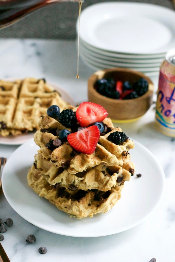A carafe of maple syrup being poured on a stack of waffles topped with blueberries, strawberries and blackberries.