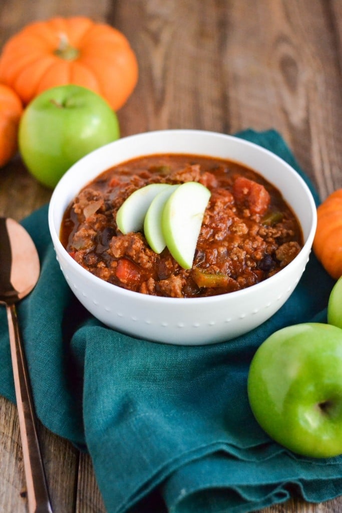A bowl of Pumpkin Apple Cider Turkey Chili topped with slices of granny smith apple and surrounded by mini pumpkins and apples.