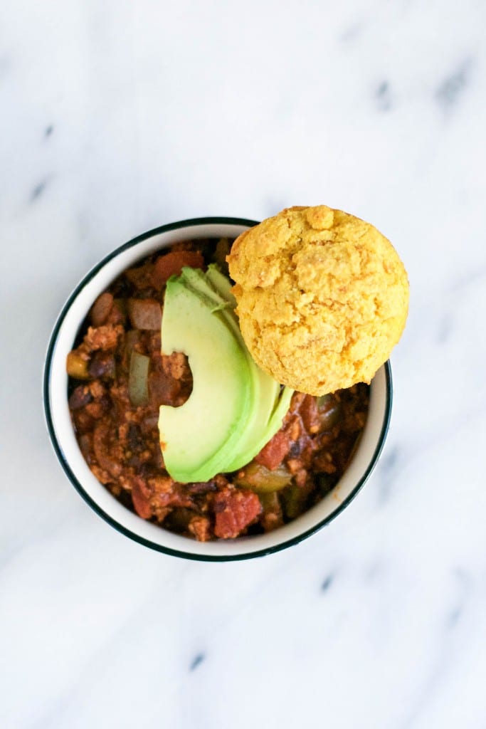 A bowl of chili topped with sliced avocado and a sweet potato cornbread muffin. 