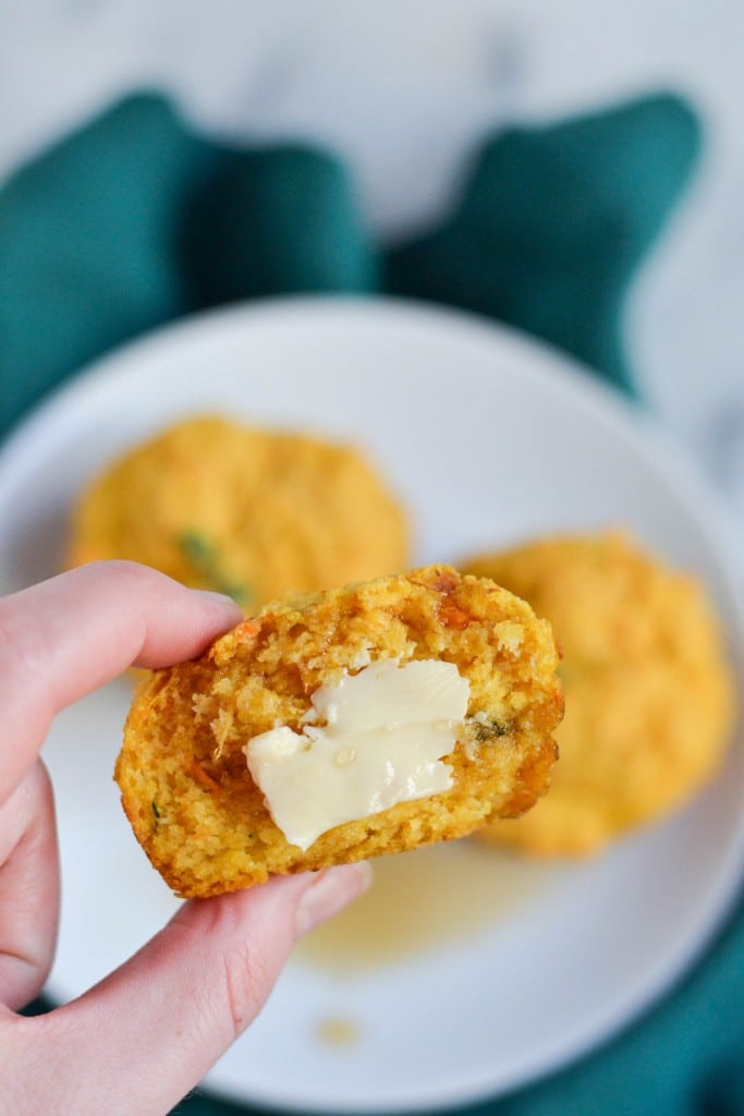 A hand holding a piece of a sweet potato cornbread muffin spreaded with butter.