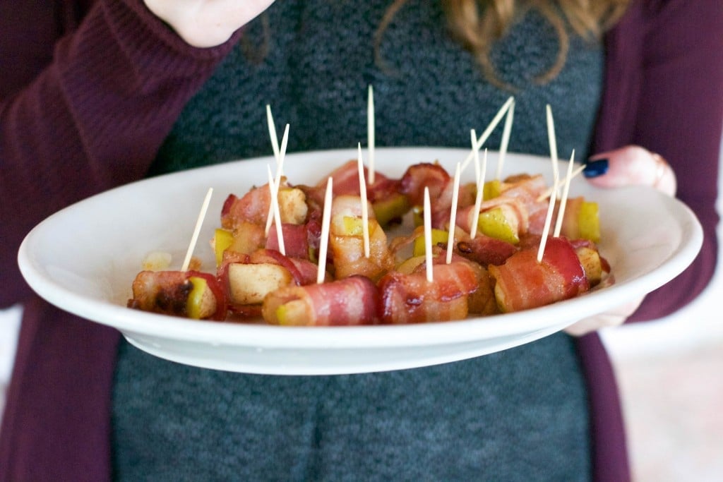A woman holding a platter of bacon wrapped apples with toothpicks sticking out of them. 