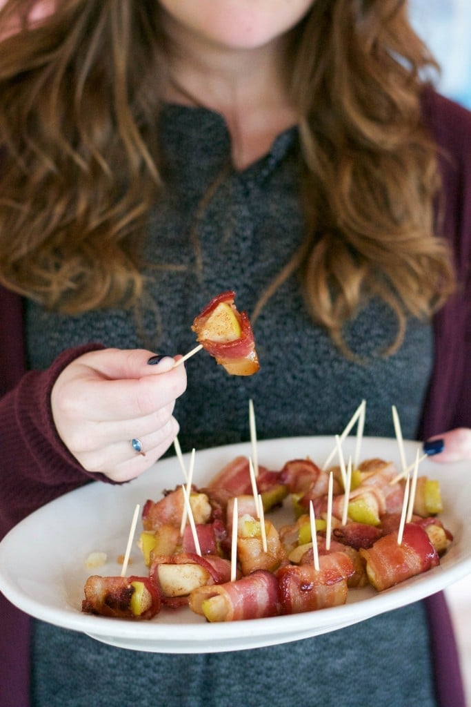 A woman holding a platter of bacon wrapped apples in one hand and picking up a piece with the other. 