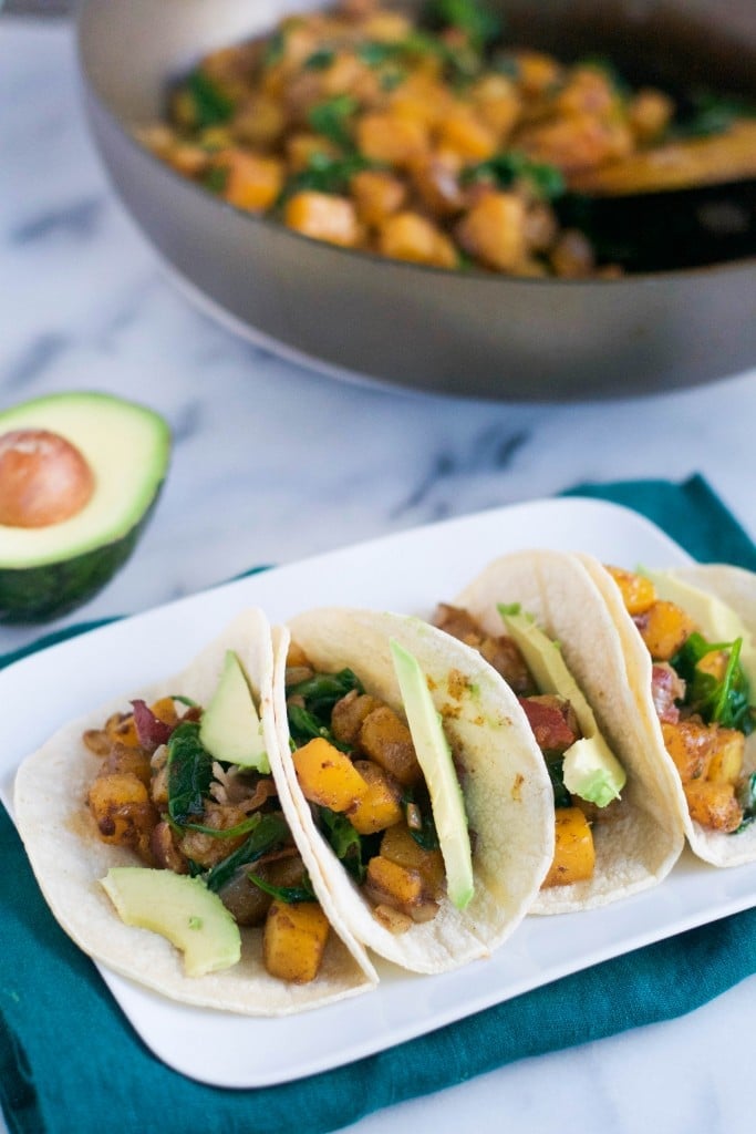 A platter of four butternut squash tacos with avocado and a skillet in the background.