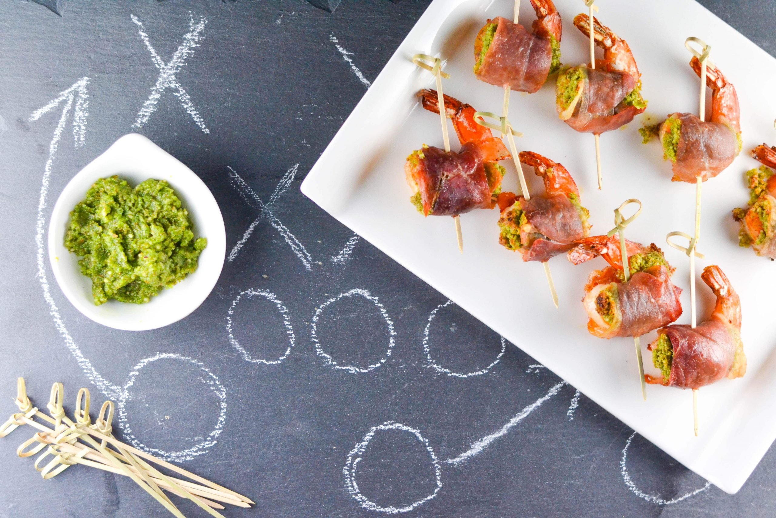 A platter of Prosciutto Wrapped Pesto Shrimp with a small bowl of pesto next to it and some cocktail sticks. 