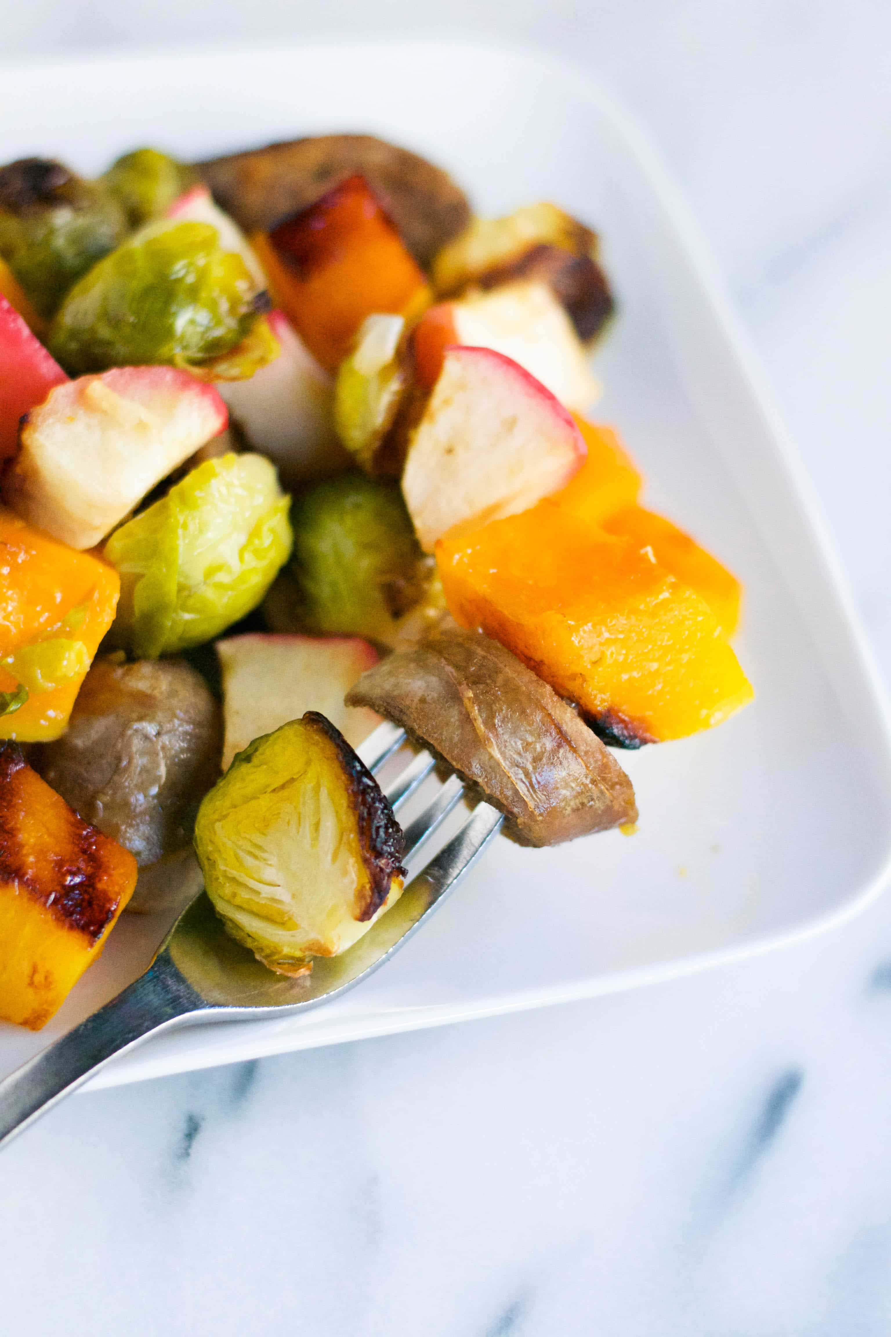 A plate of sausage, brussels sprouts, butternut squash, and apples with a fork on it. 