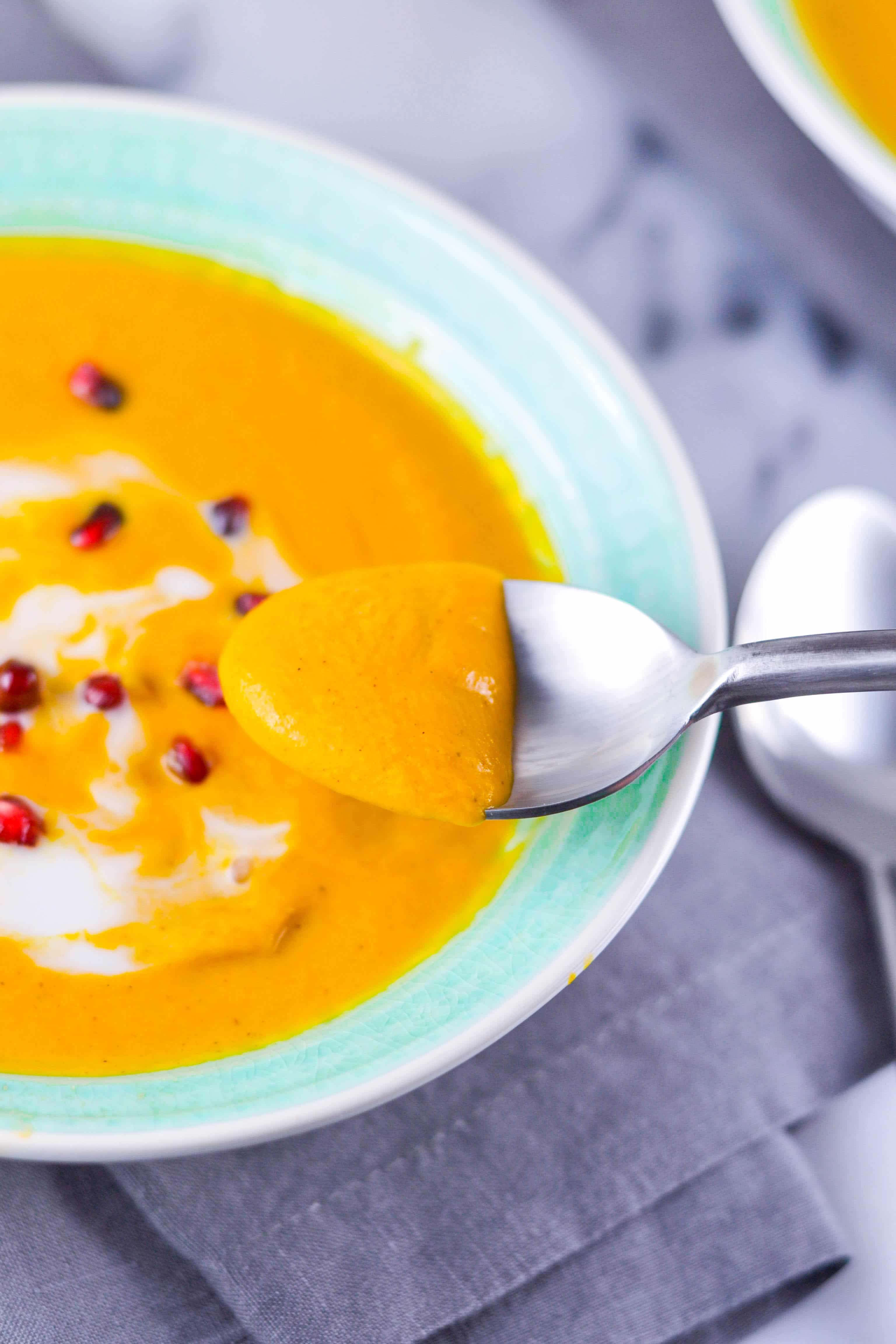 A spoon with carrot soup in it in front of a bowl of carrot soup with coconut milk swirl and pomegranate arils. 