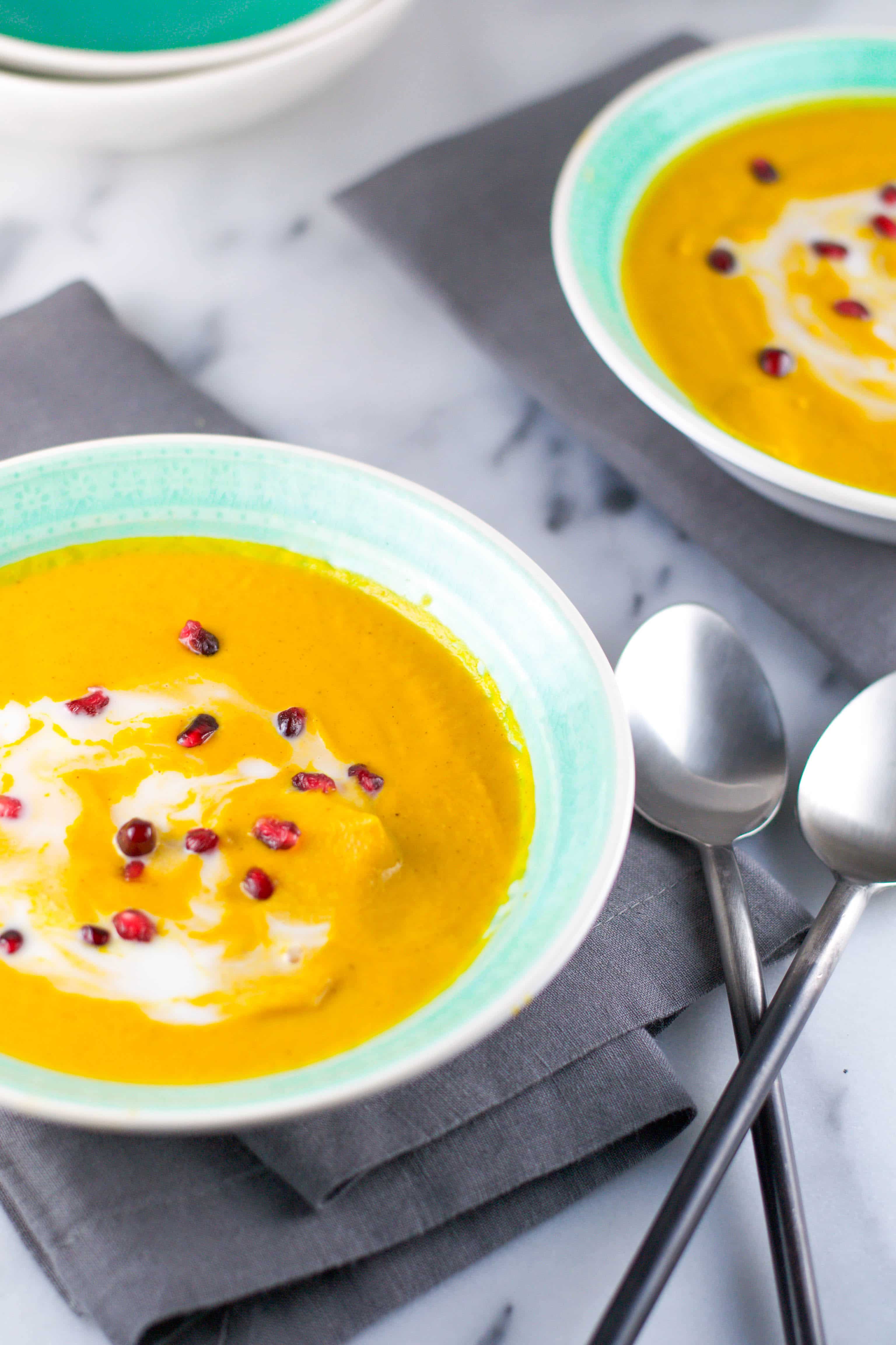 Turmeric Carrot Soup topped with coconut milk and pomegranate arils in a bowl with two spoons. 