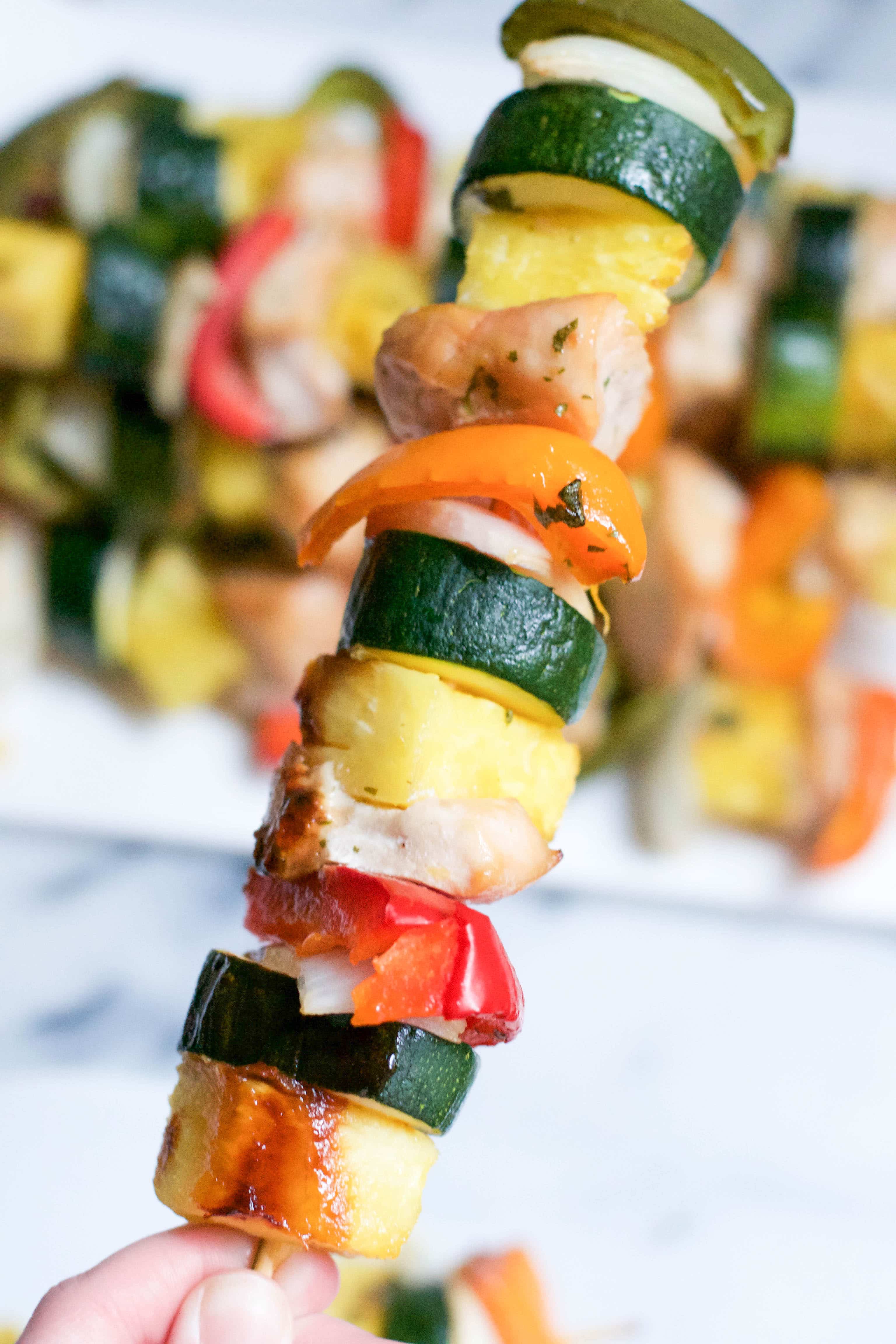 A hand holding a chicken kabob skewer with zucchini, pineapple and bell peppers. 