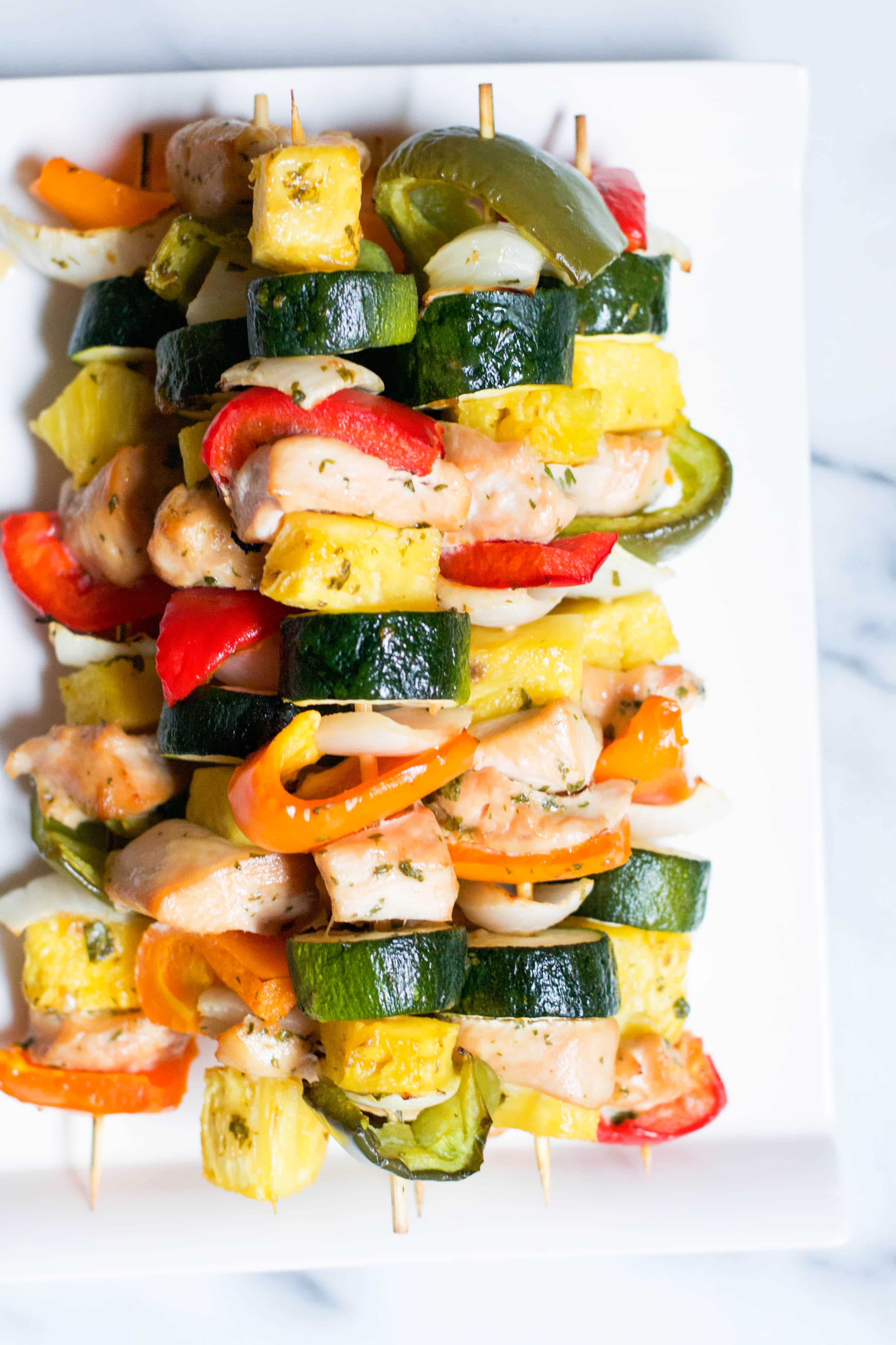 A pile of Baked Honey Lime Chicken Kabobs on skewers with zucchini, pineapple, and bell peppers on a platter.