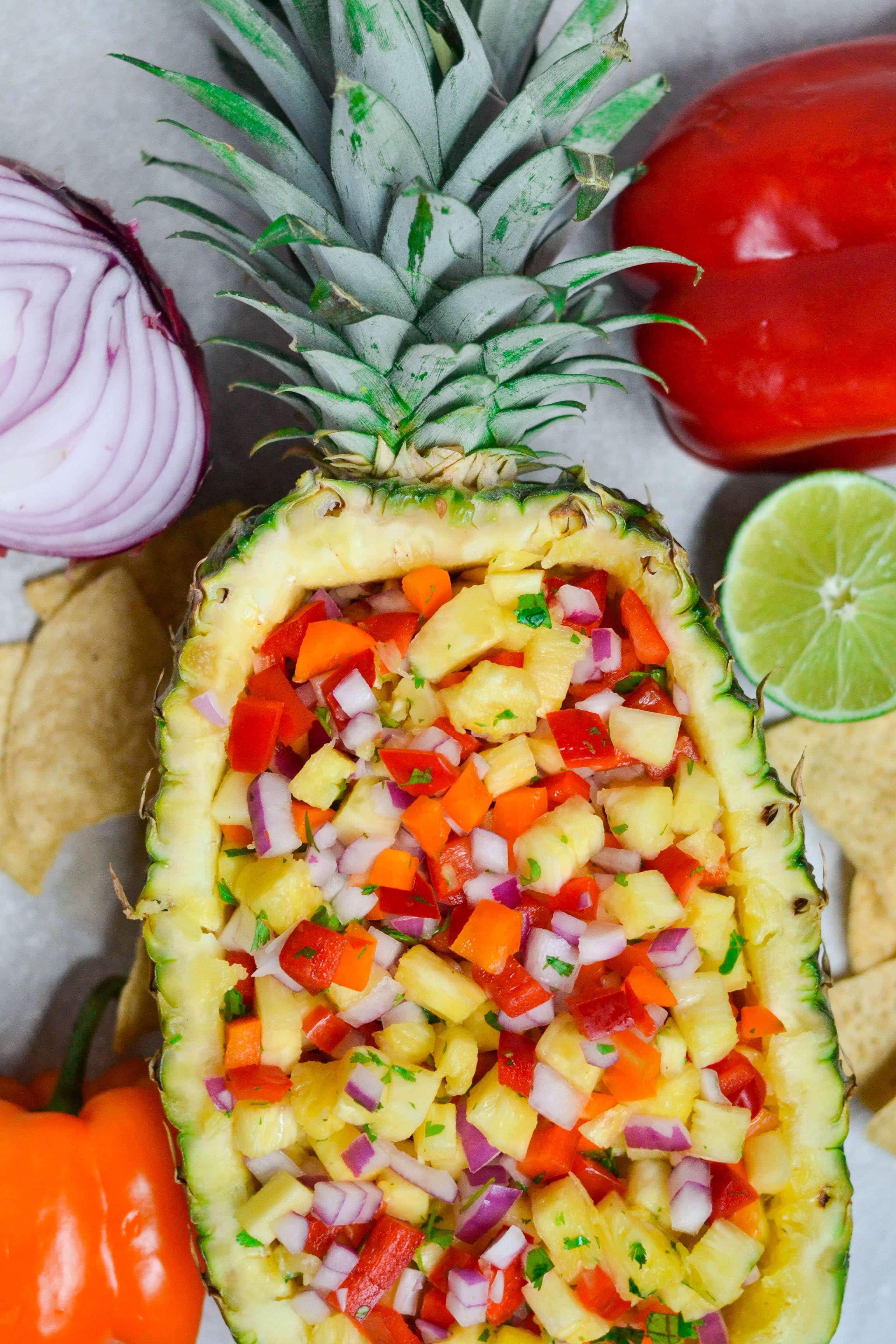 A pineapple boat filled with pineapple salsa with limes, bell peppers, tortilla chips and red onion on the table. 