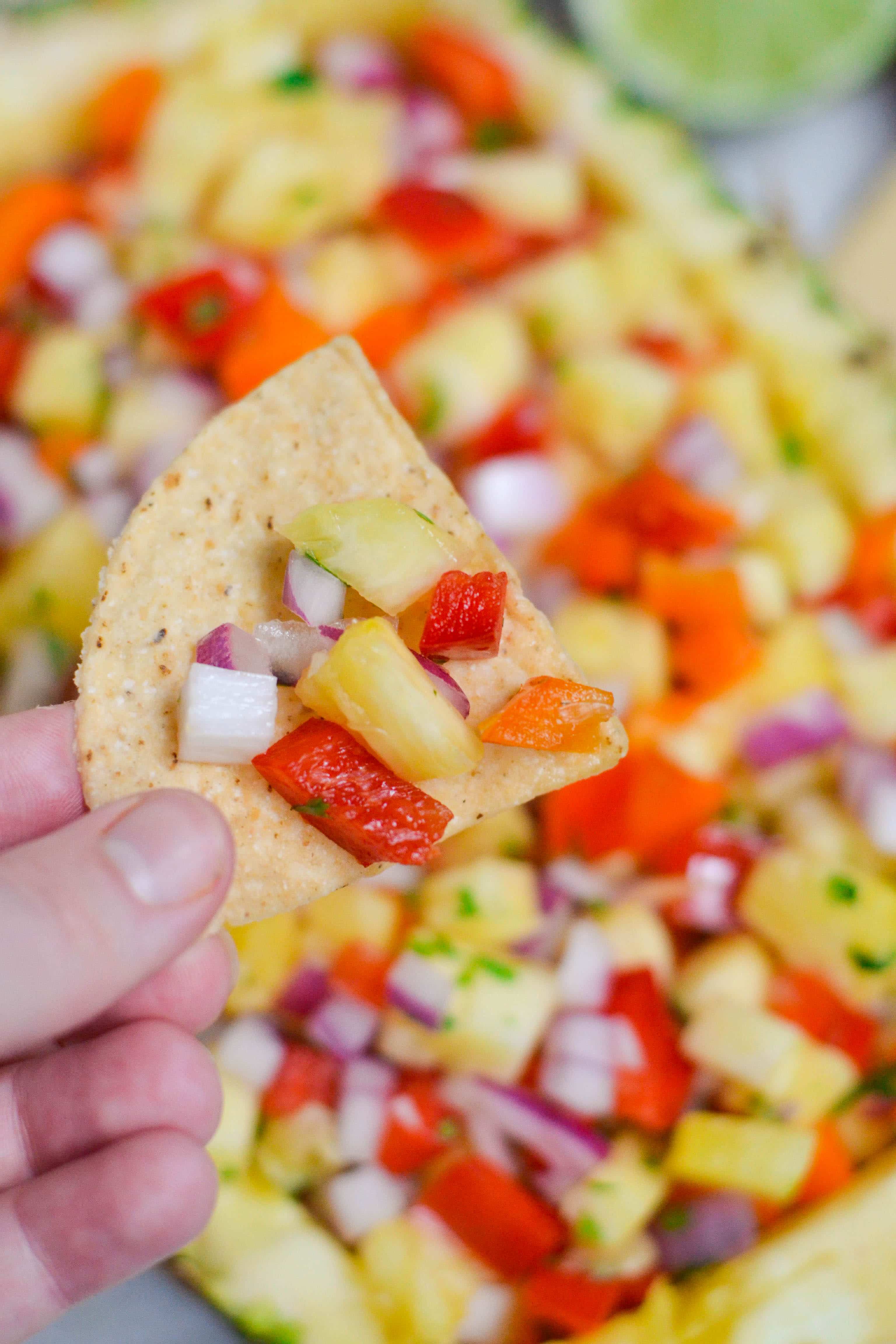 A hand holding a tortilla chip filled with pineapple salsa with a bowl of pineapple salsa below it. 
