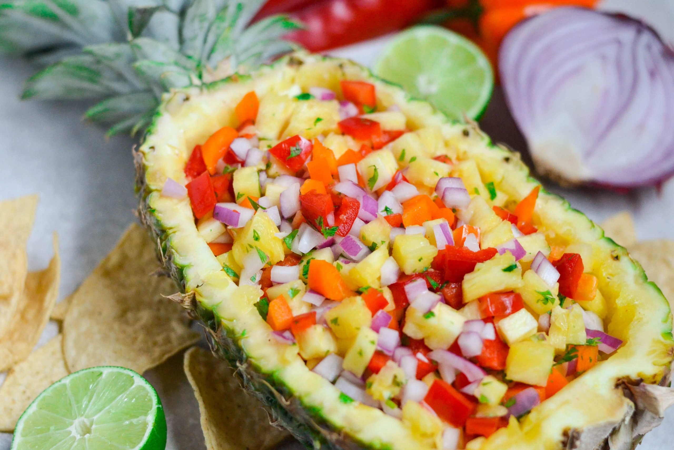 A pineapple boat filled with pineapple salsa with lime, tortilla chips, red onions in the background on the table. 
