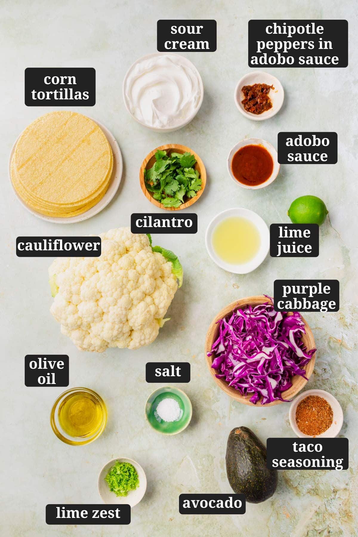 An overhead view of small bowls of ingredients to make cauliflower tacos on a green marble surface with text overlays over each ingredient.