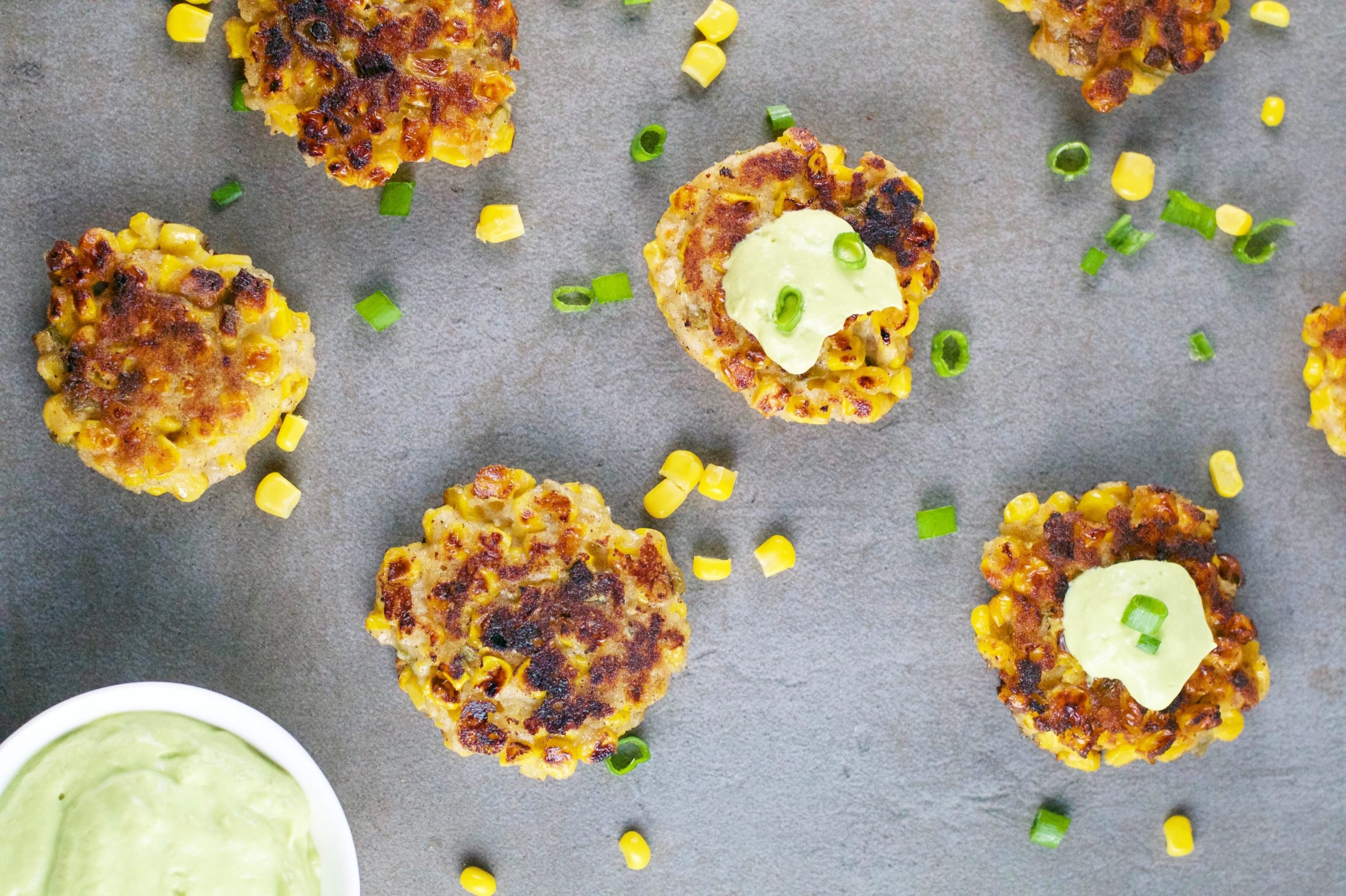 An overhead view of corn fritters on a table with a bowl of avocado crema and corn and green onions scattered on the table. 