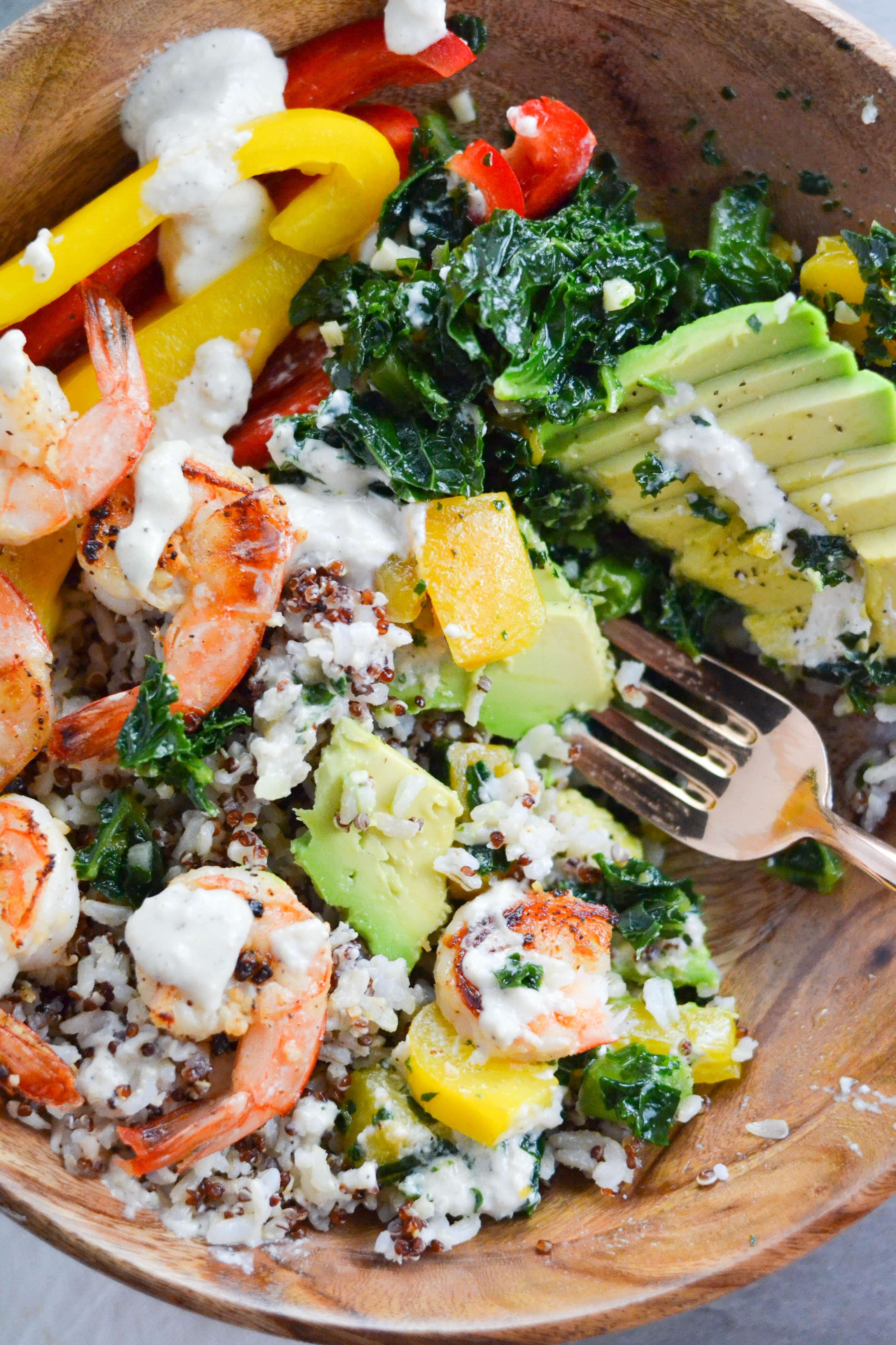 A bowl with shrimp, butternut squash, kale, avocado, quinoa, rice, bell peppers and creamy cashew sauce with a fork in it. 