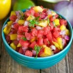 A turquoise bowl filled with vibrant watermelon salsa—diced watermelon, mango, red onion, and cilantro—sits on a wooden surface with chips, a yellow pepper, a lime, and a slice of watermelon in the background.