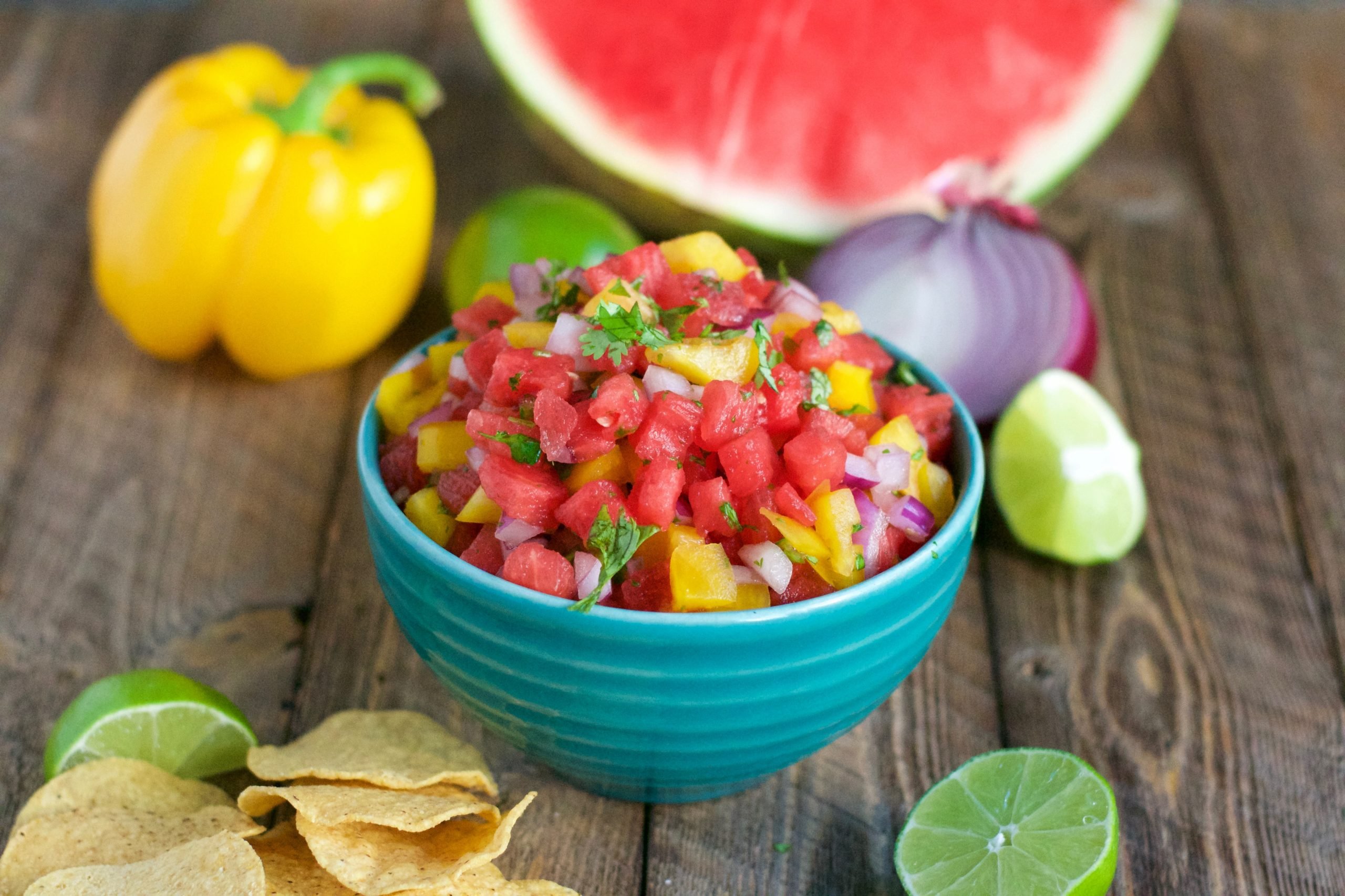 A bowl of watermelon salsa on a wood table with tortilla chips, yellow bell pepper, red onion and limes on the table. 