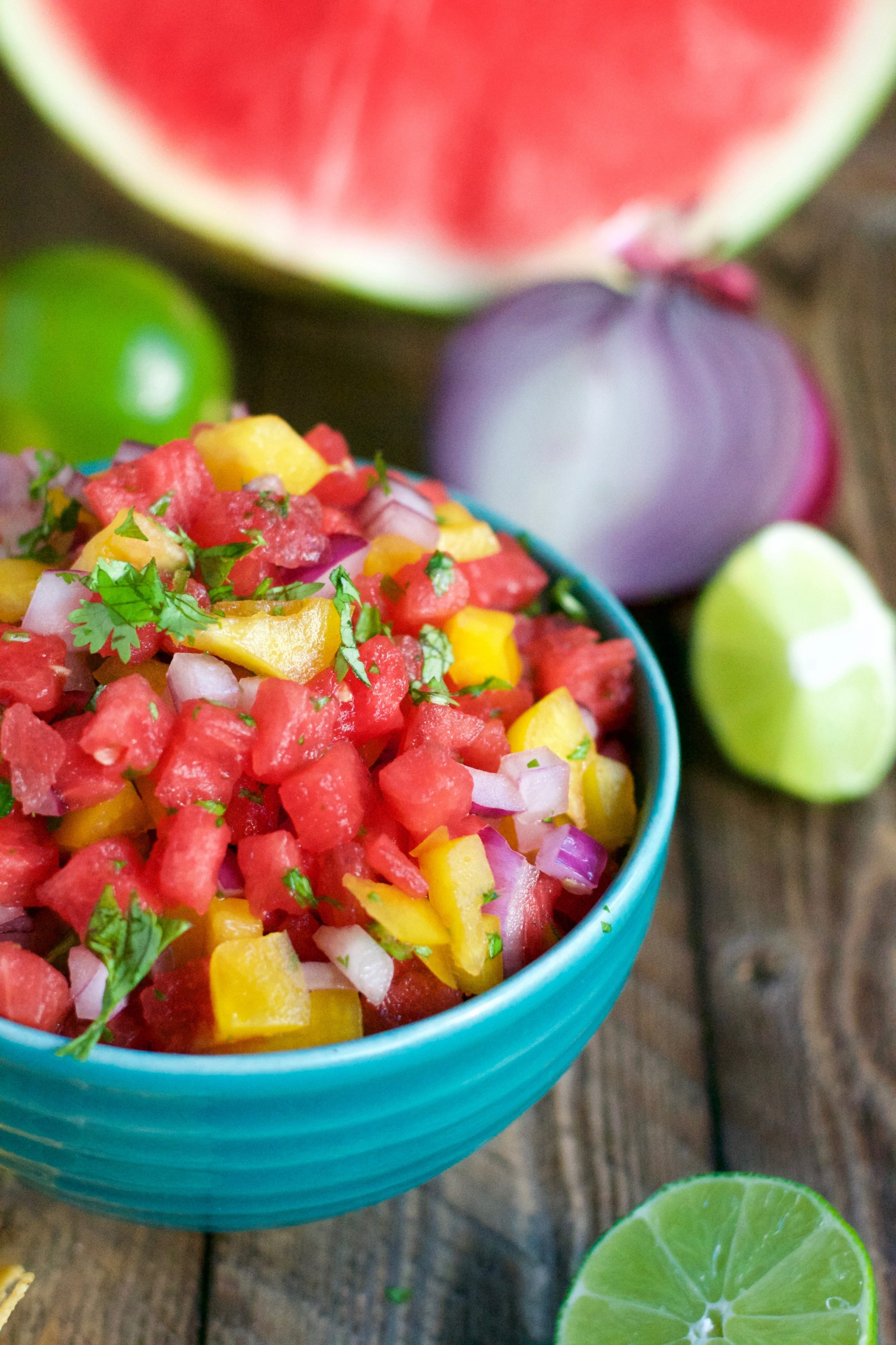 A turquoise bowl with watermelon salsa topped with cilantro on a wooden table with limes, red onion and watermelon 