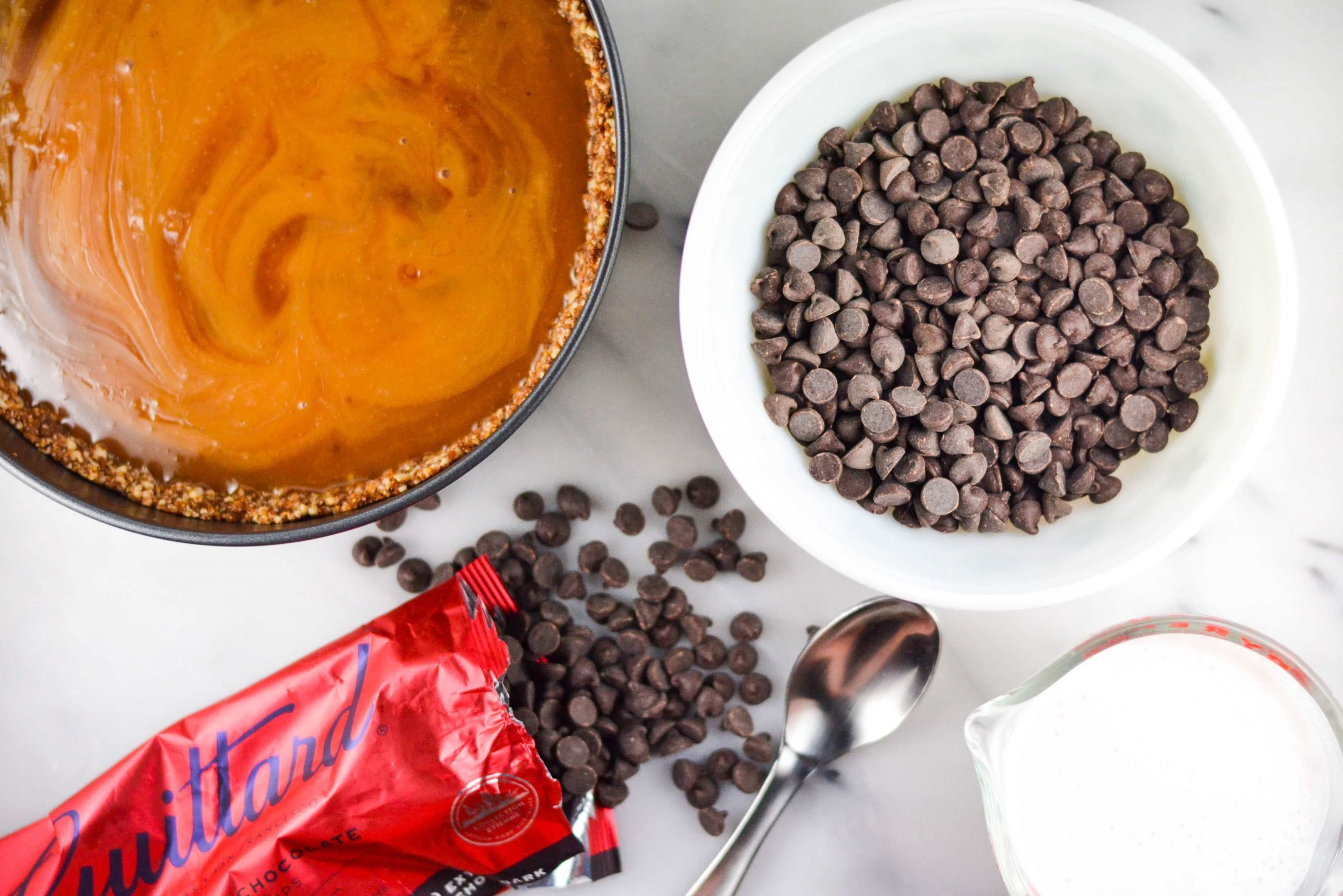 A springform pan with caramel sauce on top of a nut crust, a bowl of chocolate chips and a ramekin of heavy cream. 