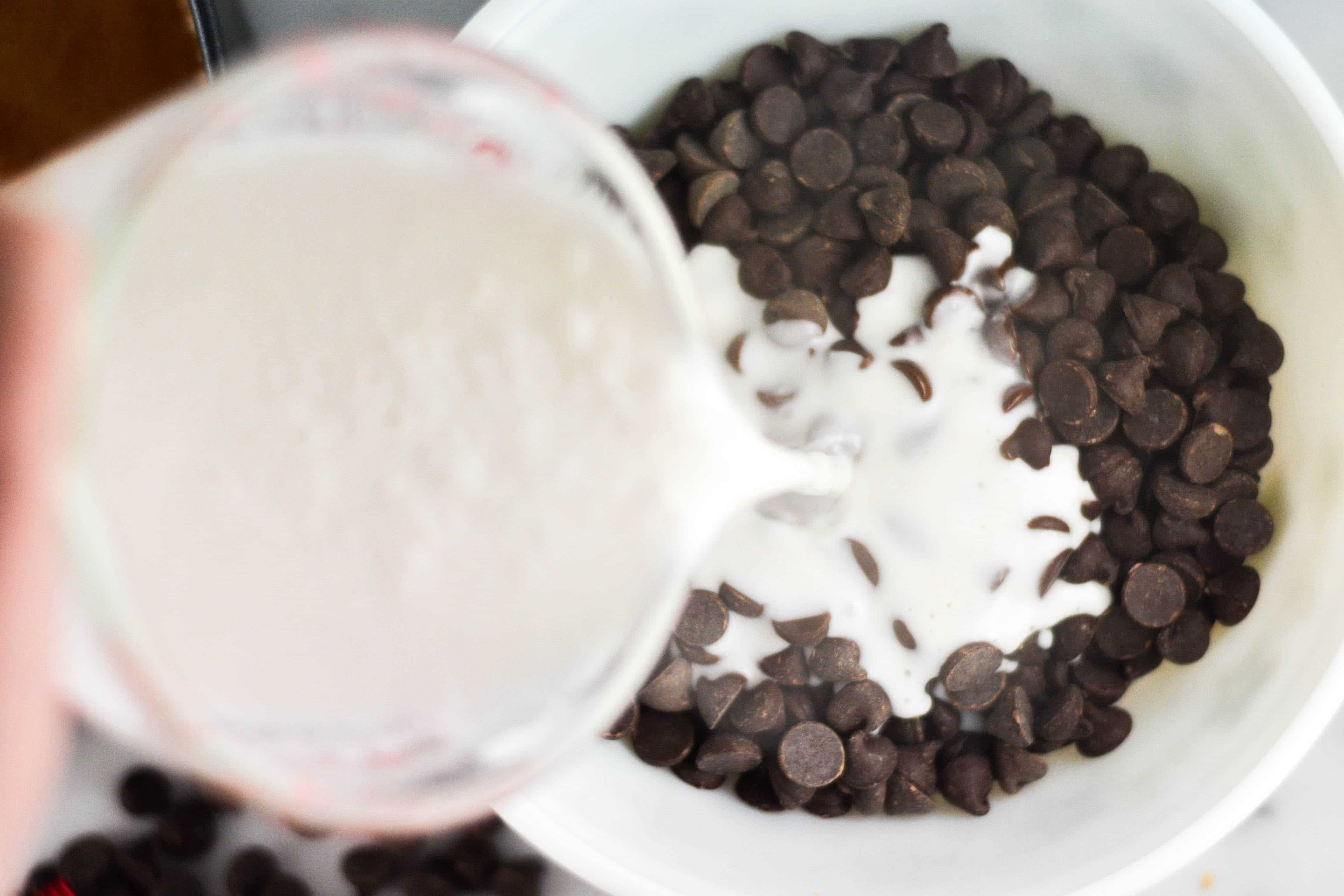 A hand pouring a measuring cup of heavy cream into a bowl of chocolate chips. 