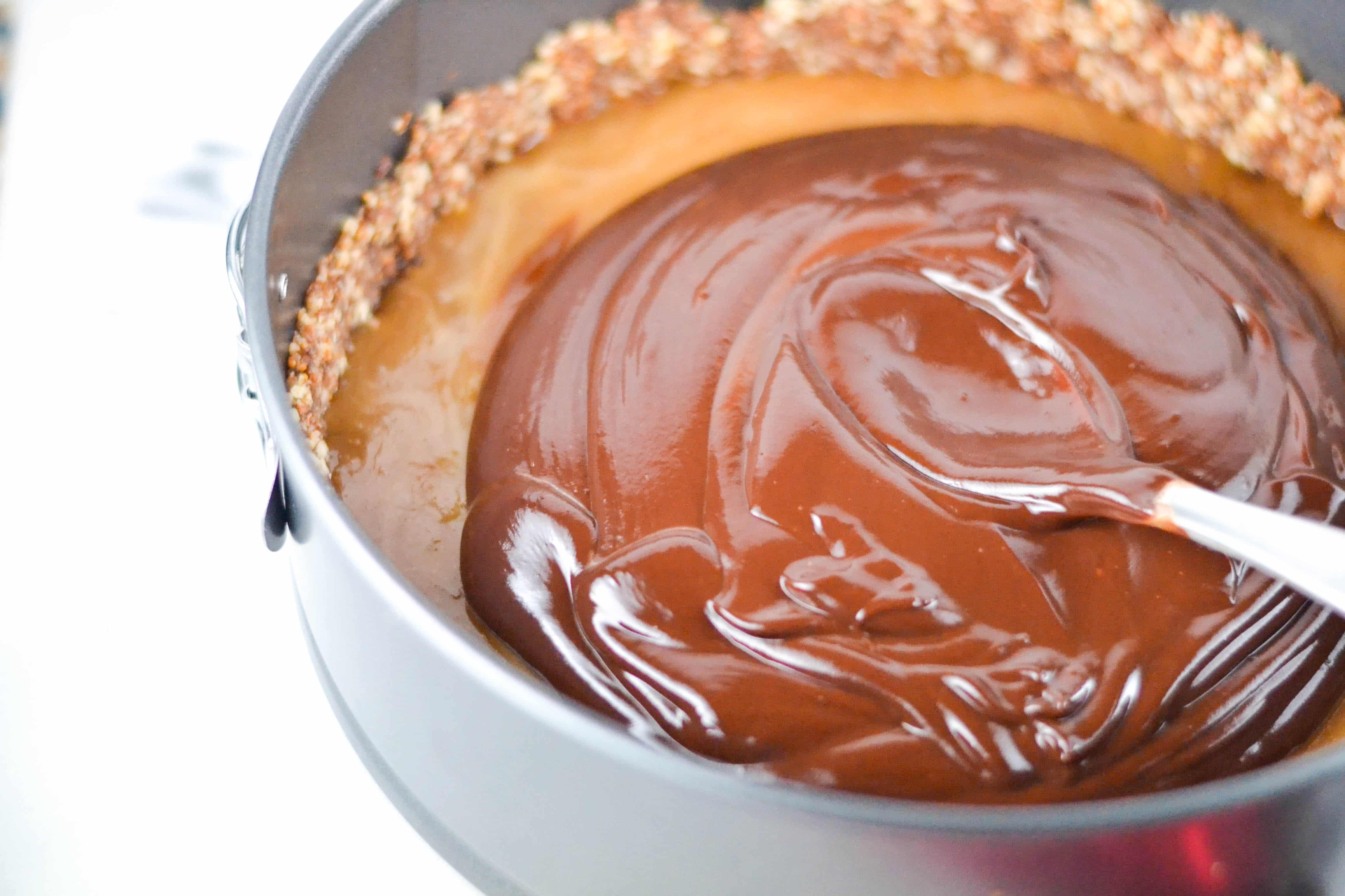 Chocolate ganache being spread with a spoon over a caramel filling and nut crust in a springform pan.  