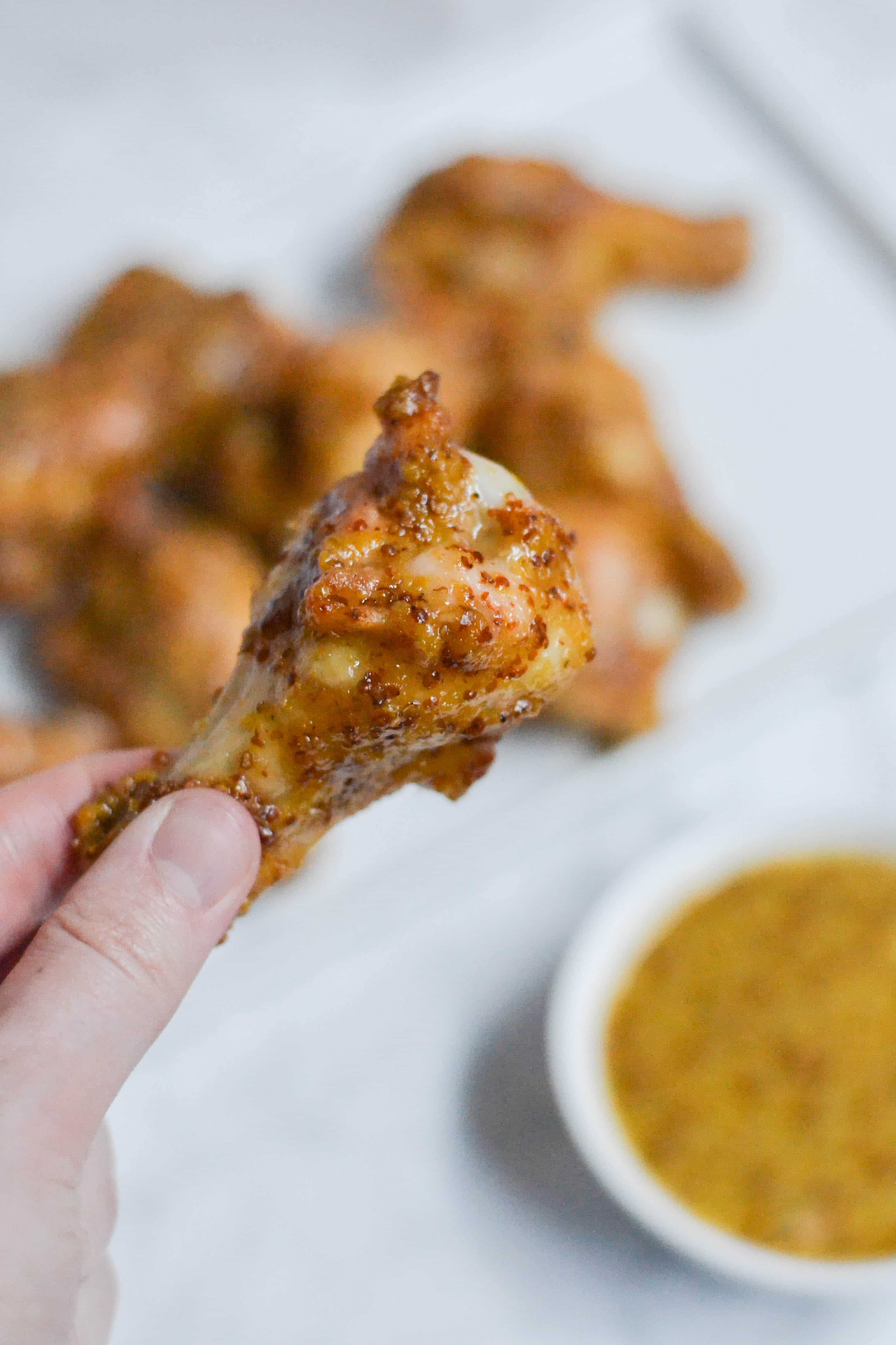 A hand holding a honey mustard chicken wing with a ramekin of honey mustard sauce in the background. 