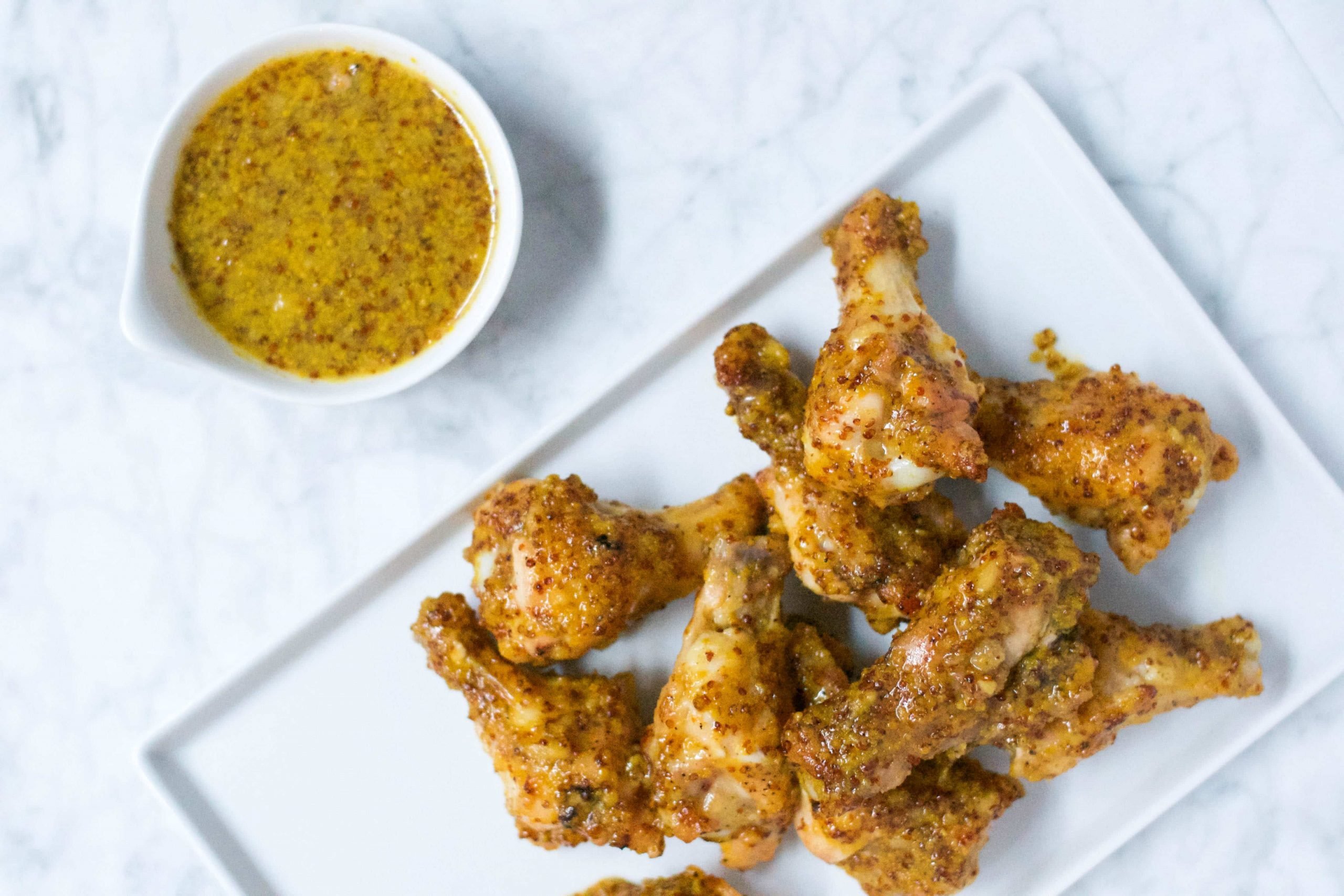 A white platter with a pile of honey mustard chicken wings and a ramekin of honey mustard sauce on the side. 