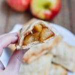 A hand holding a half-eaten gluten-free vegan apple hand pie with a visible spiced filling, with whole apples and more pies on a plate in the background.