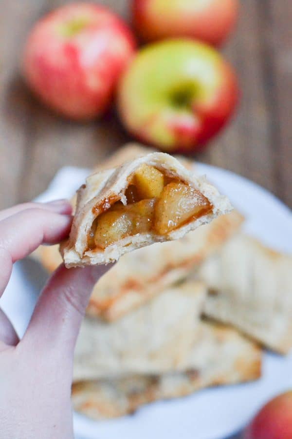 A hand holding a half-eaten gluten-free vegan apple hand pie with a visible spiced filling, with whole apples and more pies on a plate in the background.