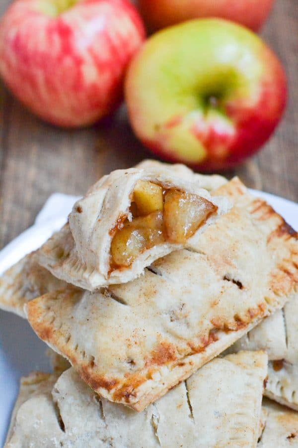A stack of gluten-free vegan apple hand pies, one broken open to show a spiced apple filling, with fresh apples in the background.