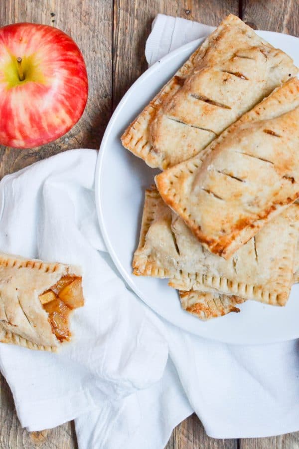 A plate of gluten-free vegan apple hand pies next to a fresh apple and one partially eaten pastry.