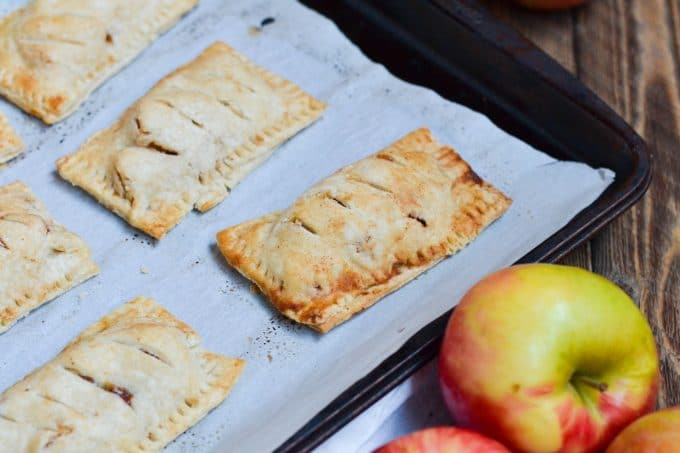 Gluten-free vegan apple hand pies on a parchment-lined baking sheet next to fresh apples.