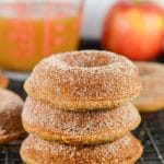Four cinnamon sugar-coated gluten-free vegan apple cider donuts are stacked on a cooling rack, with a red apple and a glass measuring cup filled with brown liquid in the blurred background.