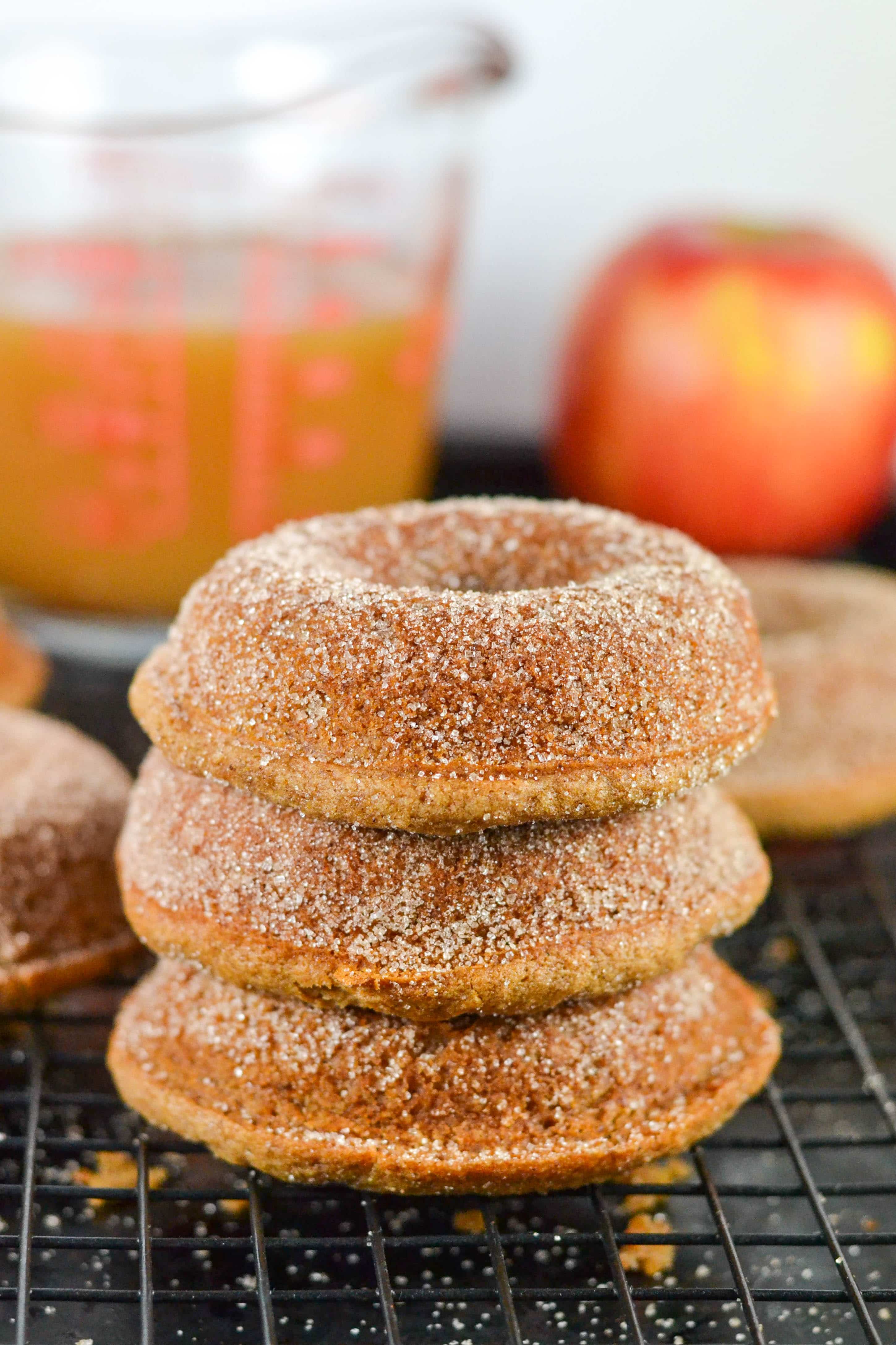 A stack of three gluten-free apple cider donuts on a cooling rack with an apple and a measuring cup of apple cider in the background. 