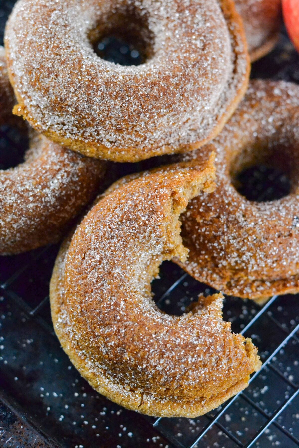 Four gluten-free vegan apple cider donuts on a cooling rack, with one donut featuring a large bite taken out of it, revealing its soft interior.