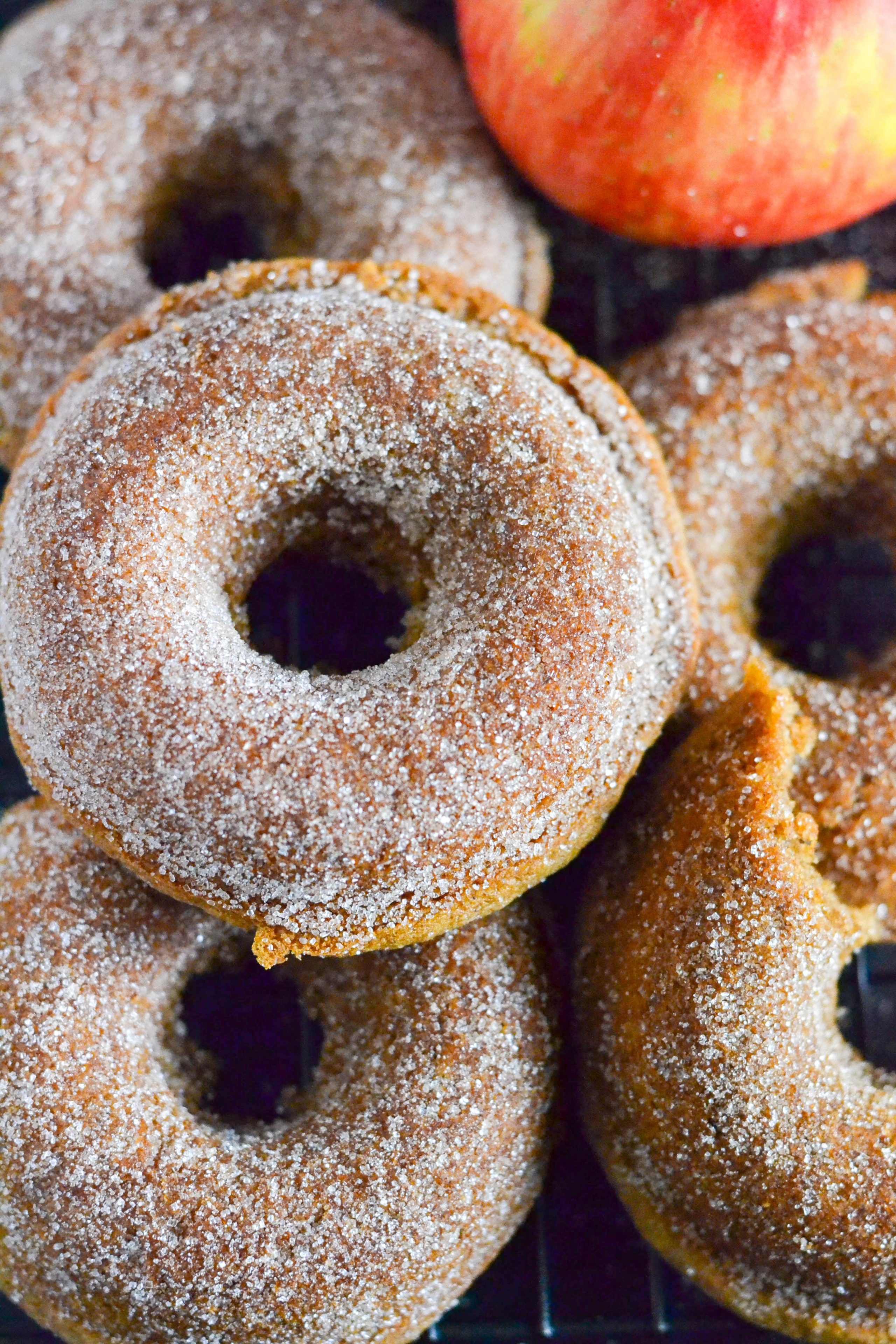 A close up of a pile of baked apple cider donuts with an apple in the background. 