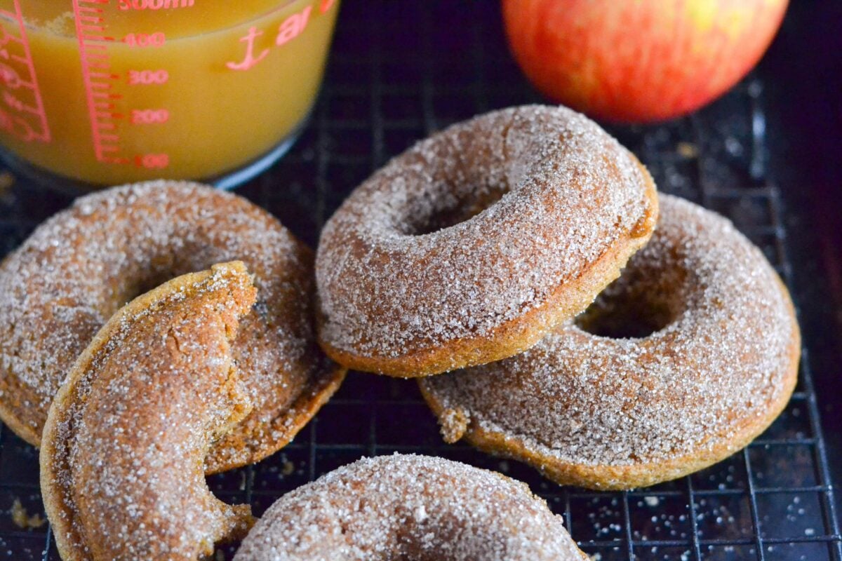 Four gluten-free vegan apple cider donuts, sugar-coated and golden, rest on a cooling rack next to a red apple and a glass measuring cup filled with apple cider. One donut has a bite taken out of it.
