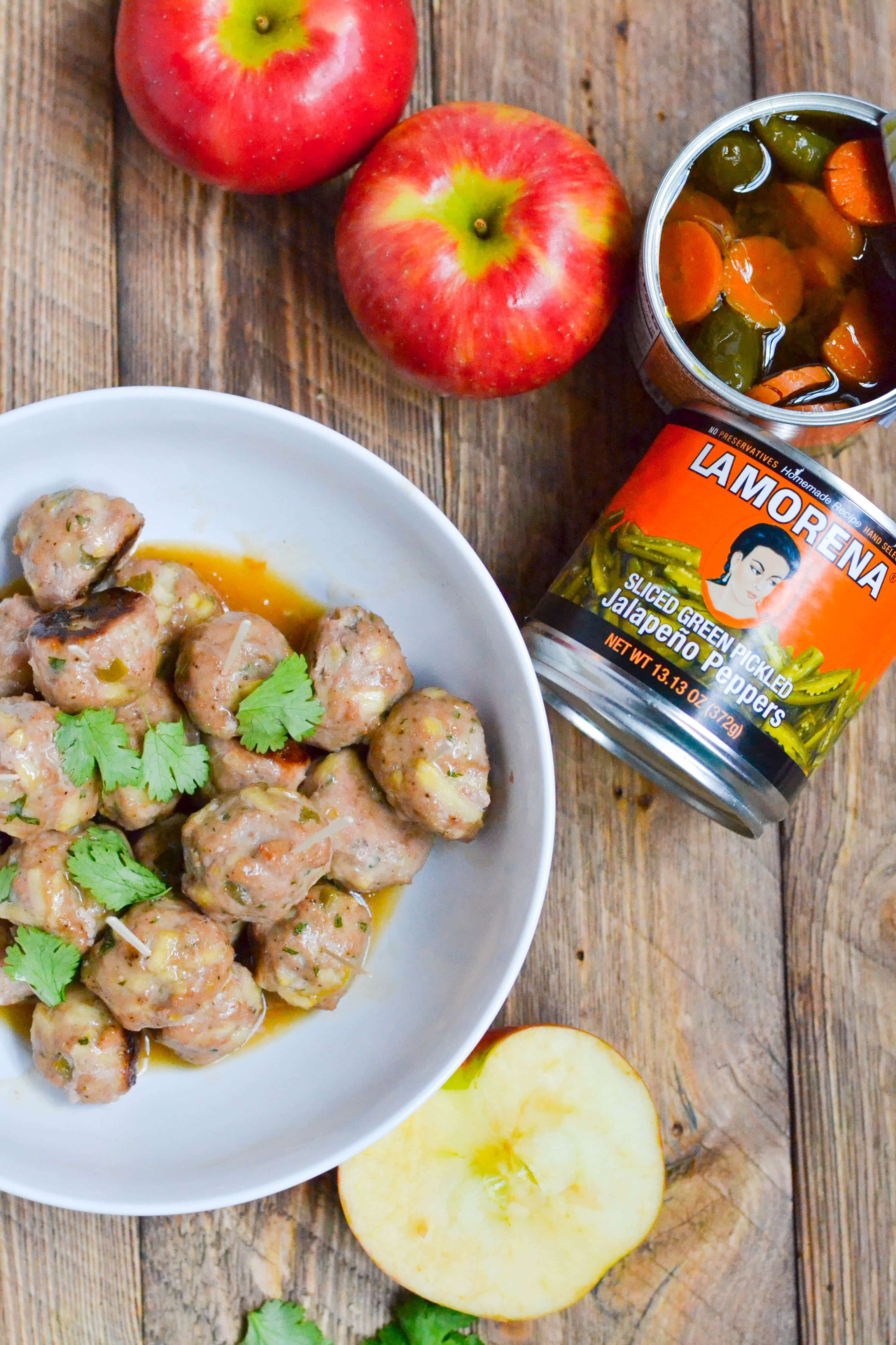 A bowl of apple meatballs with cilantro on top with apples and pickled jalapeño peppers on the table. 