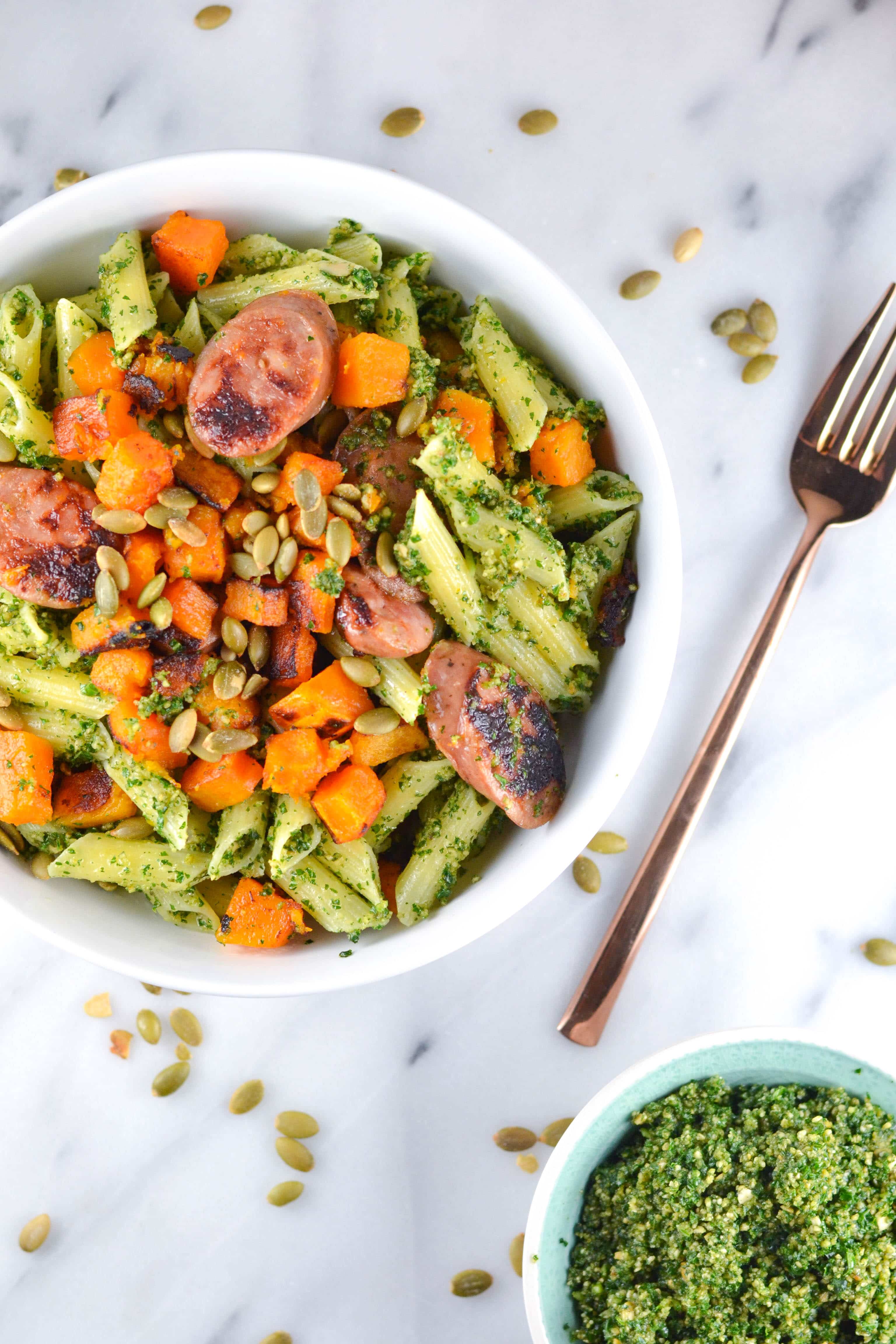A bowl of pasta with sausage, butternut squash and pepitas in it with a fork and a bowl of pesto next to it. 