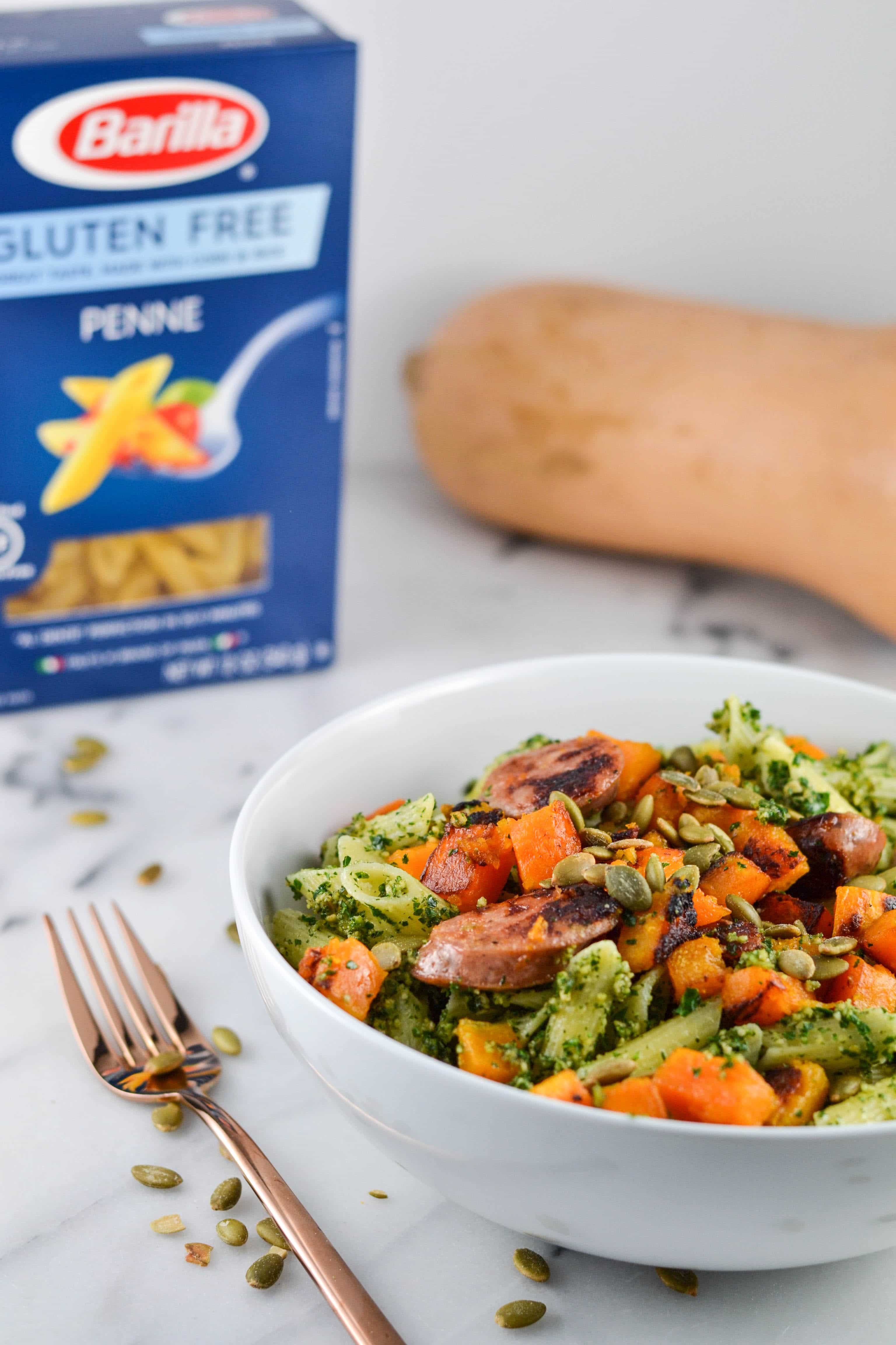 A bowl of Gluten-Free Pumpkin Seed Pesto Pasta with Butternut Squash and pumpkin seeds with a box of gluten-free pasta and butternut squash in the background. 