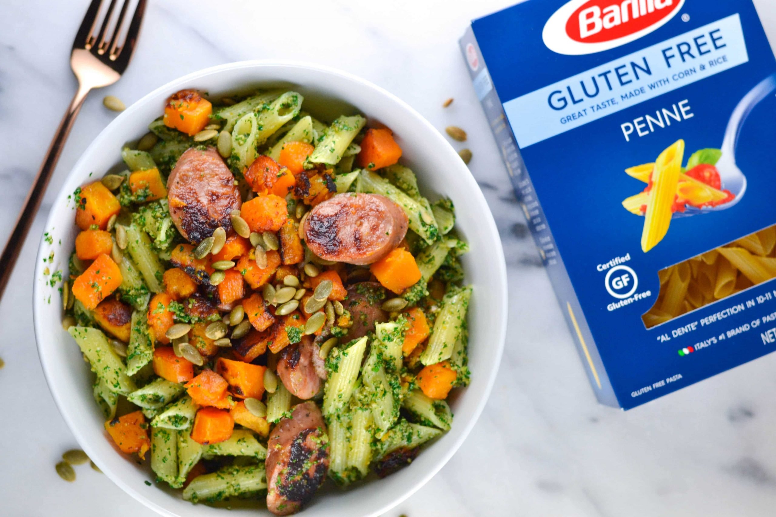 An overhead view of a bowl of pasta with pesto, sausage, butternut squash, and pumpkin seeds with a box of Barilla gluten-free penne next to it. 
