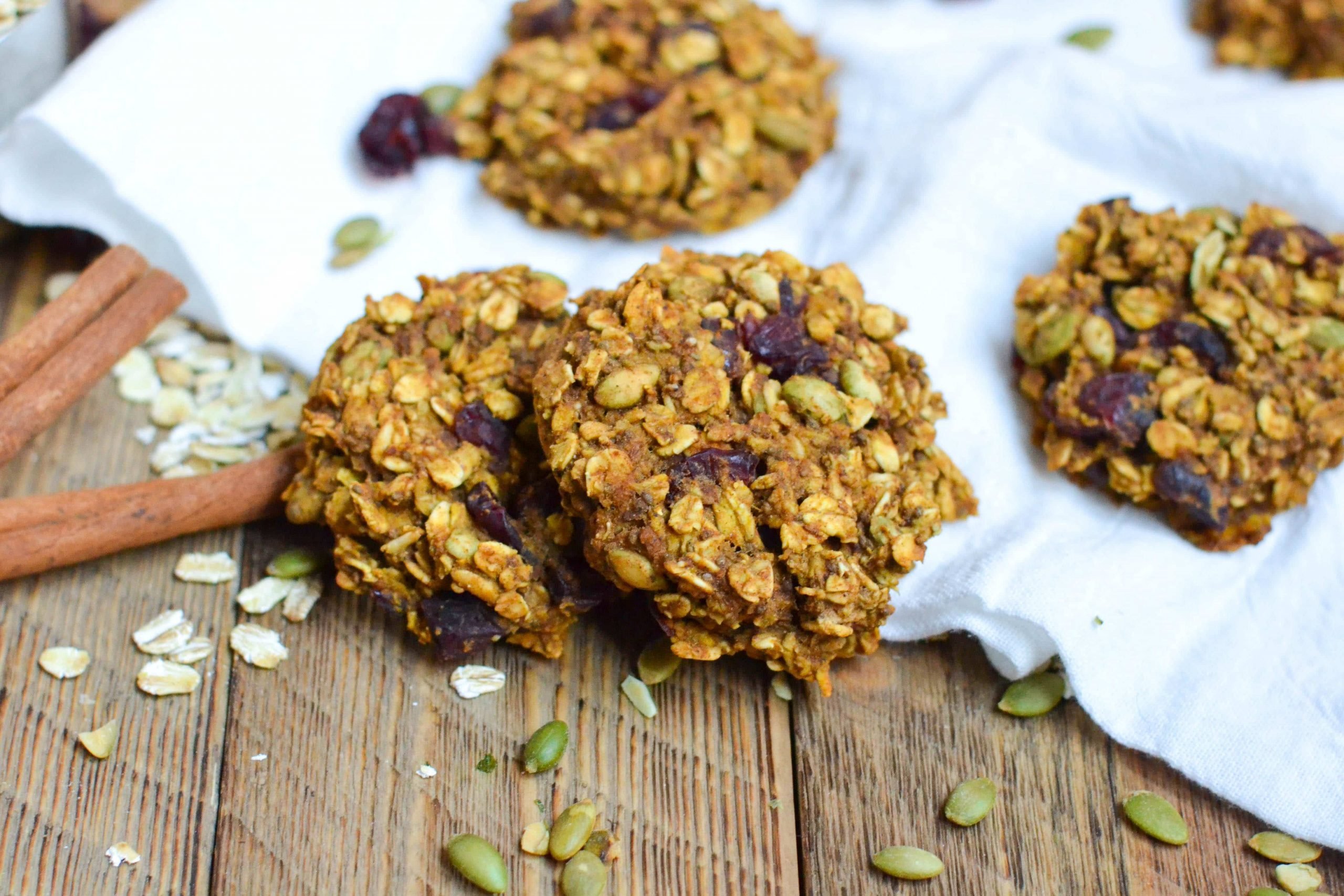 Two pumpkin breakfast cookies layered on top of each other with oats, pepitas and cinnamon sticks scattered on the surface. 