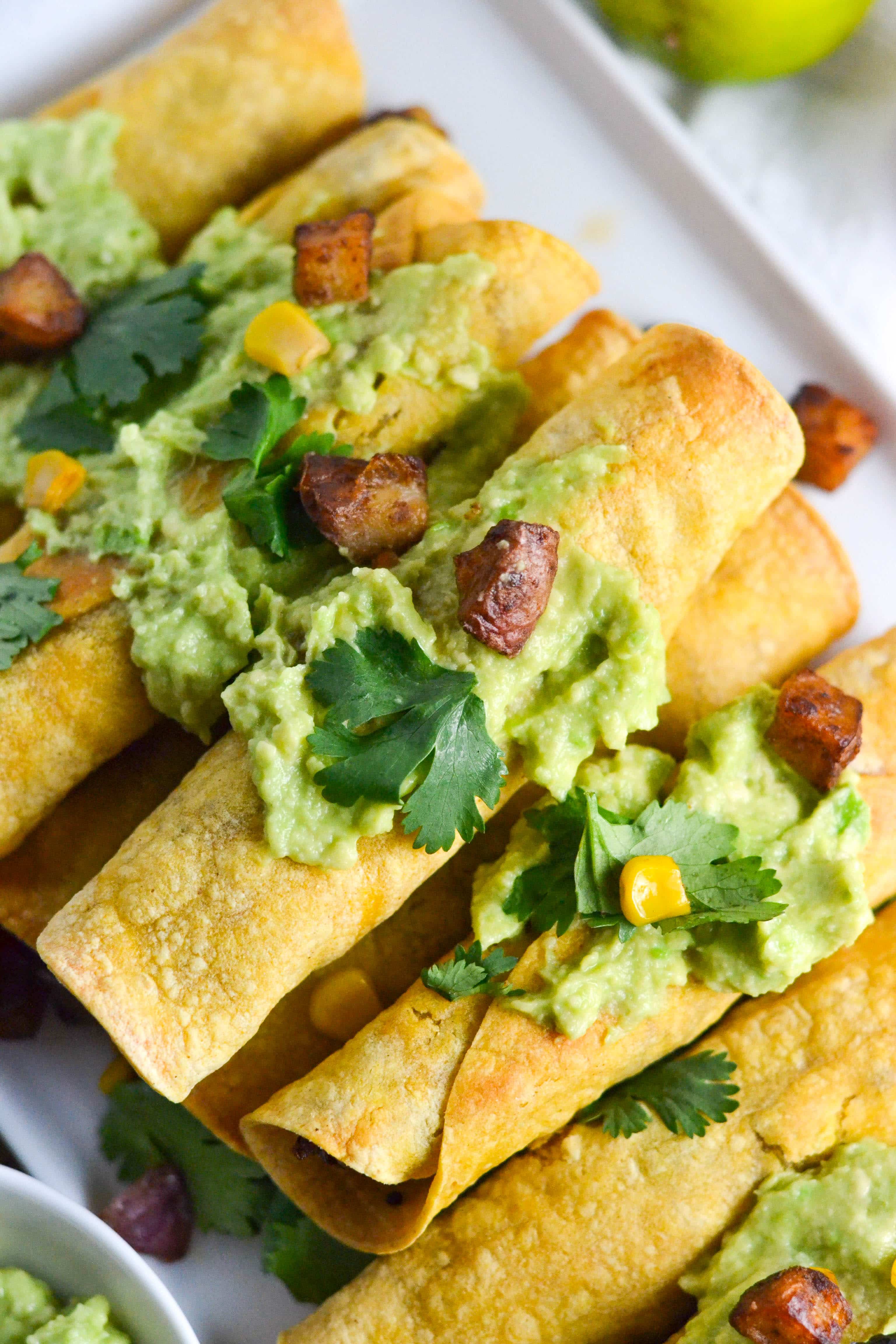 A stack of red potato taquitos topped with avocado, cilantro, corn and crispy potatoes. 