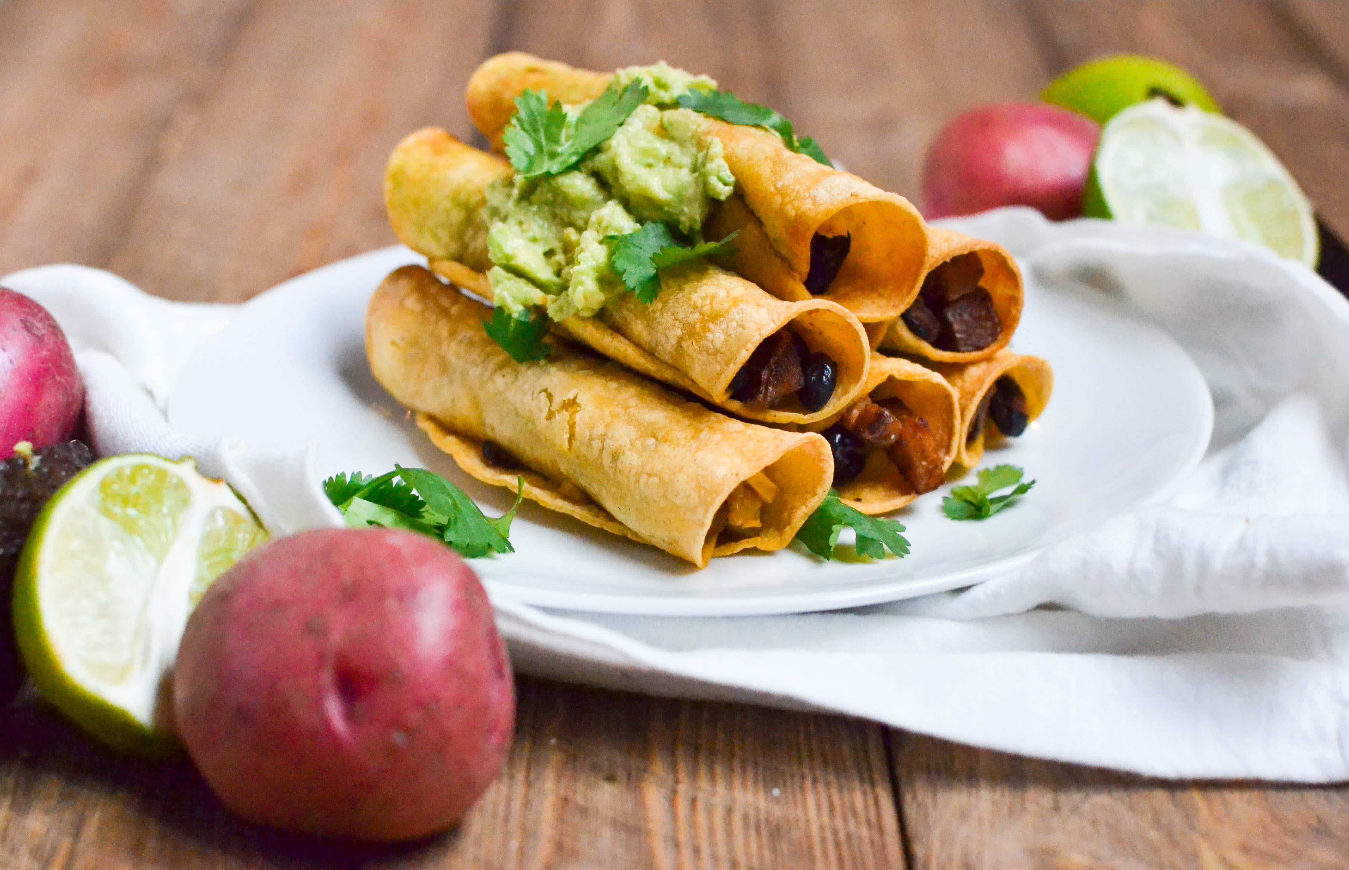 A stack of Red Potato and Black Bean Taquitos on a white plate topped with guacamole and cilantro. 