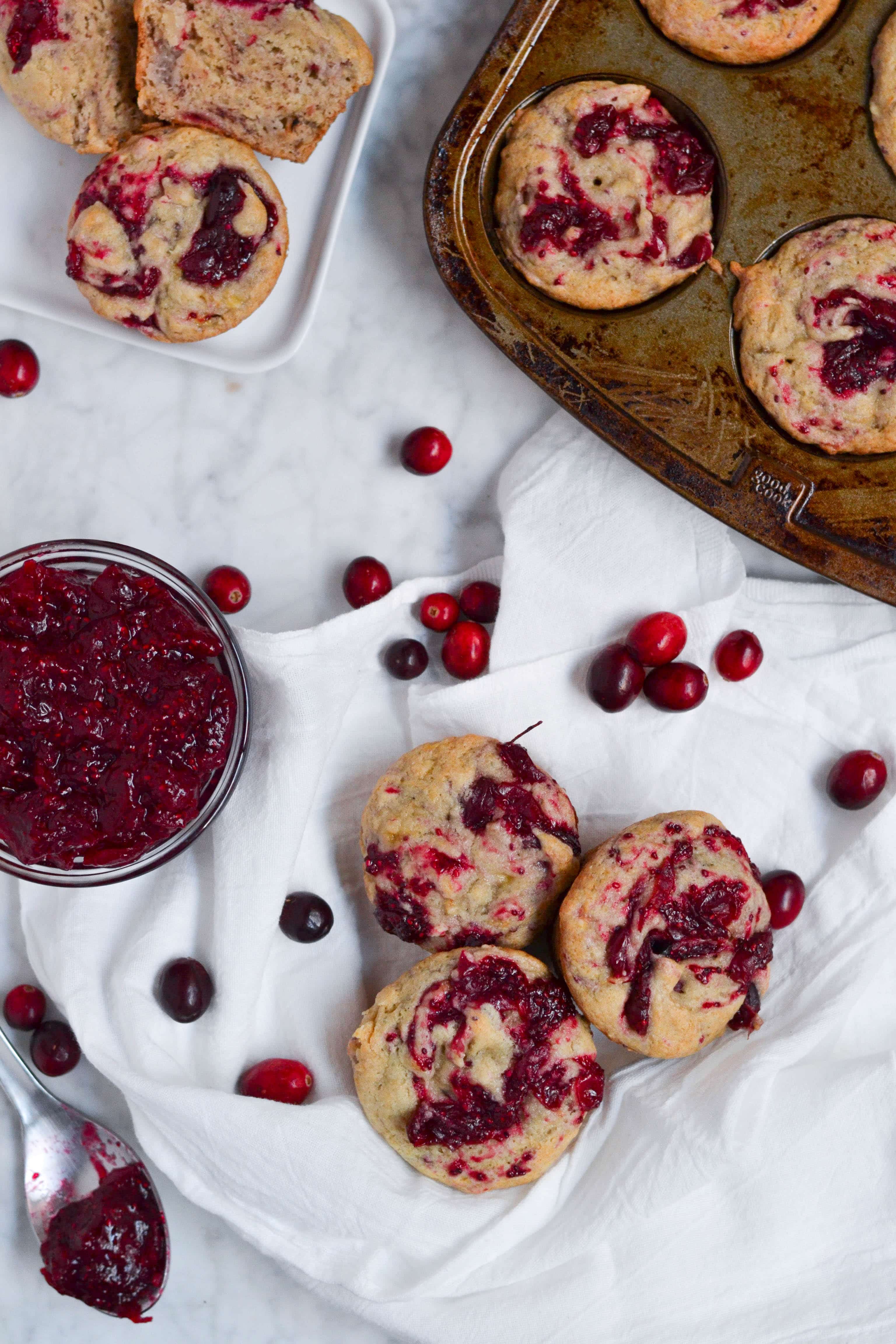 Banana Cranberry Jam Muffins on plates and in a muffin tin with a side of cranberry jam in a small bowl. 