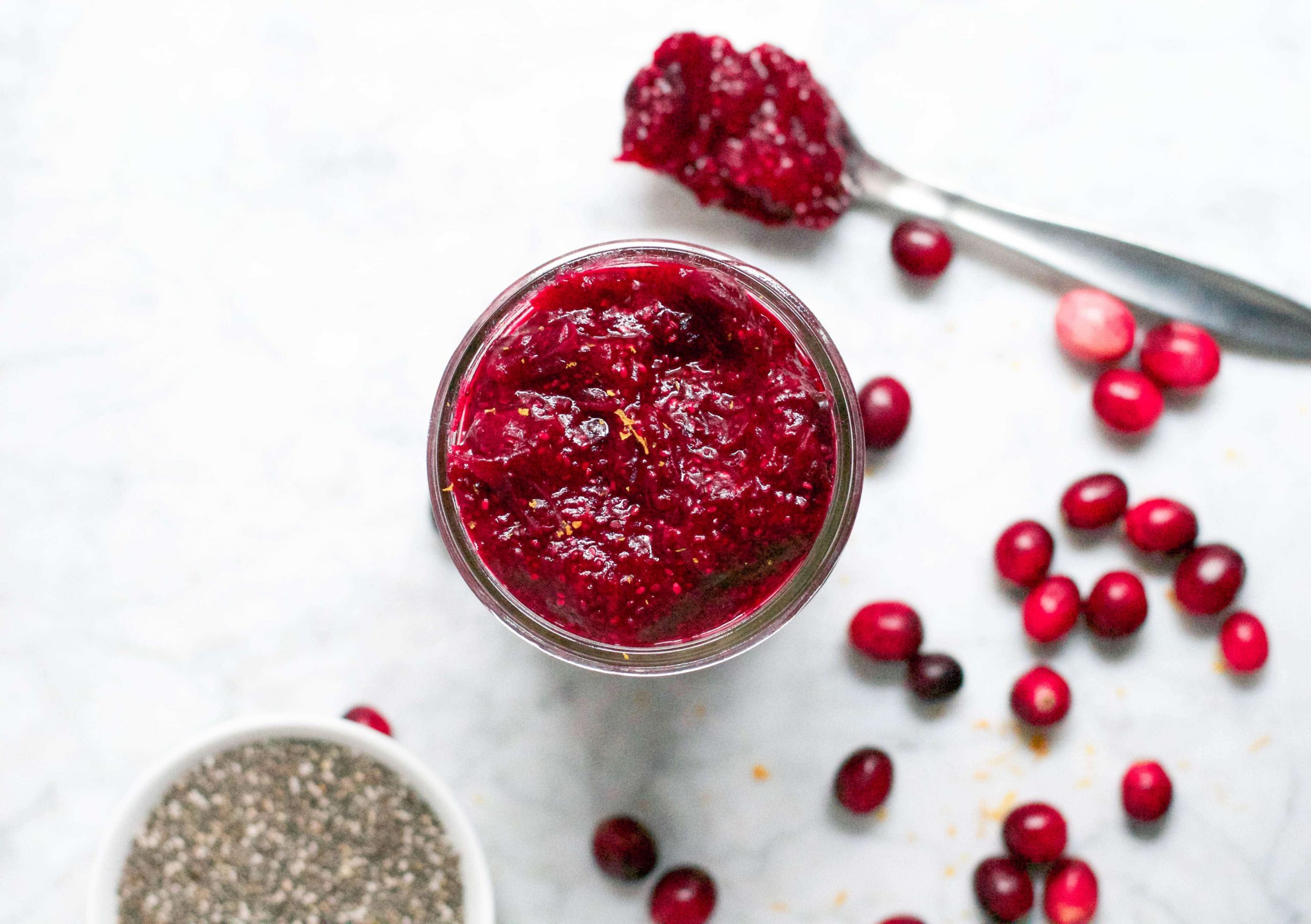 An overhead view of a jar with orange cranberry chia jam in it with fresh cranberries on the table and a bowl of chia seeds. 